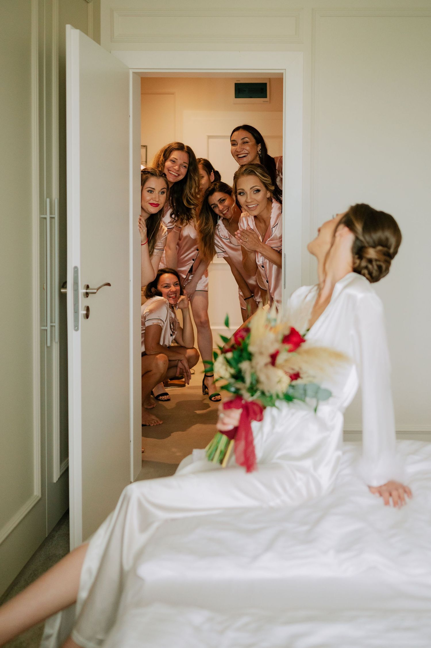 Bride in white robe greeting bridesmaids who appear in doorway during wedding morning.