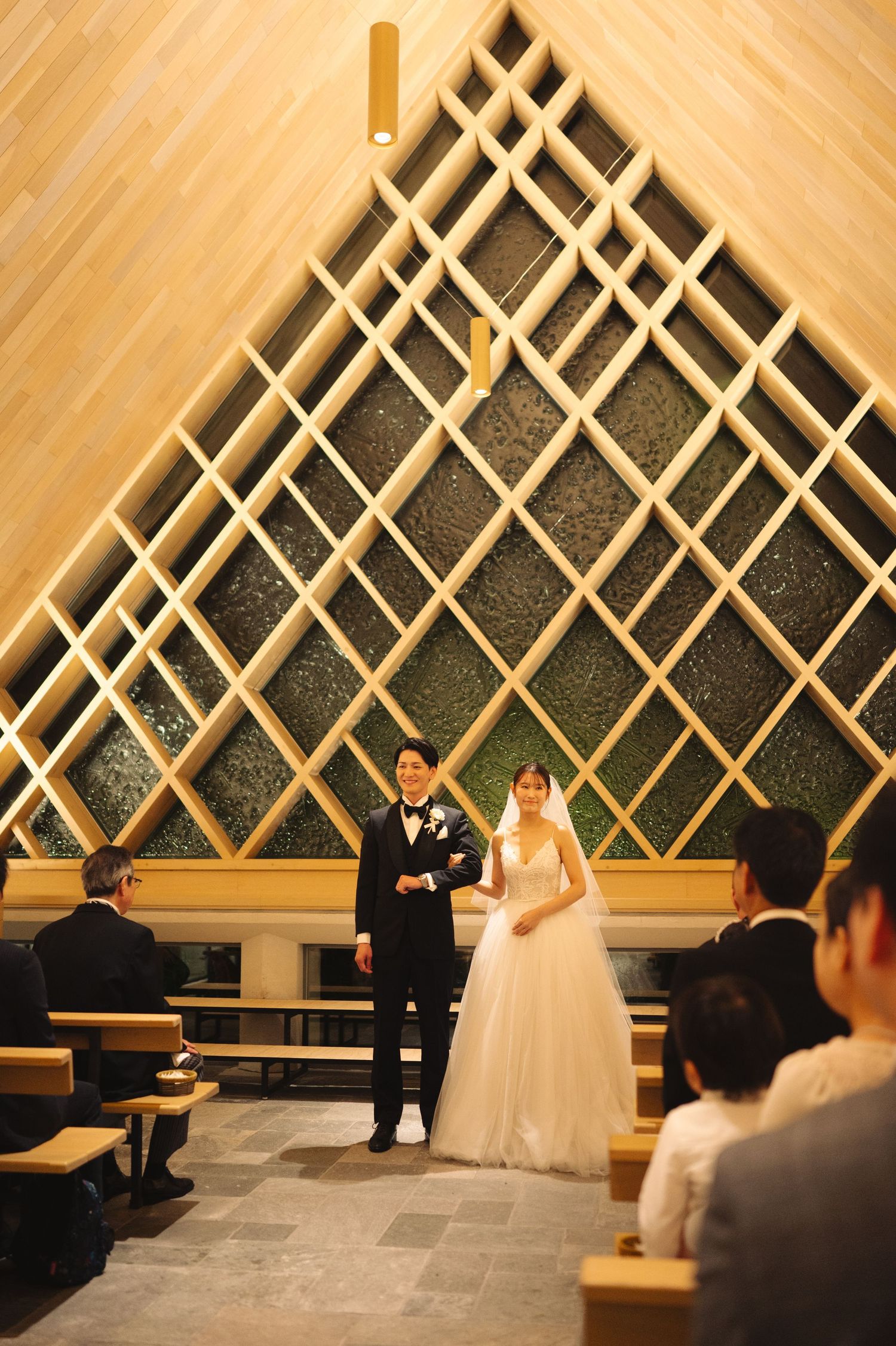 Couple stands before guests in modern chapel featuring dramatic diamond-patterned wooden wall.