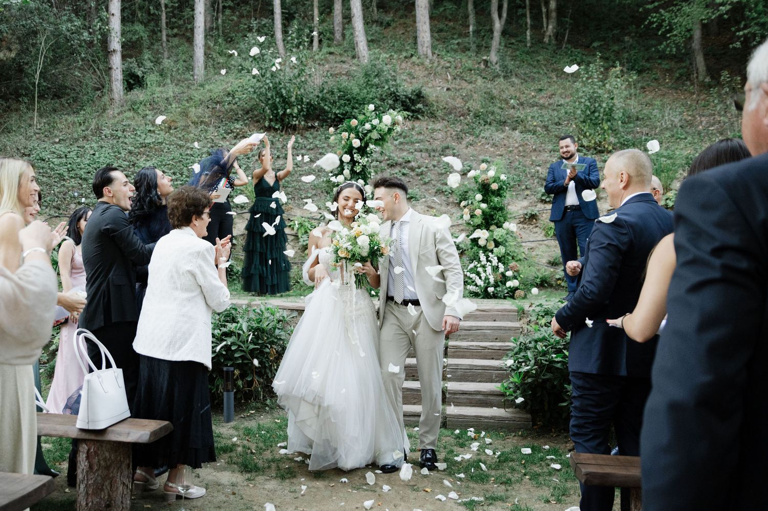 Wedding guests gather in a wooded outdoor setting with stone steps and natural greenery for a ceremony.