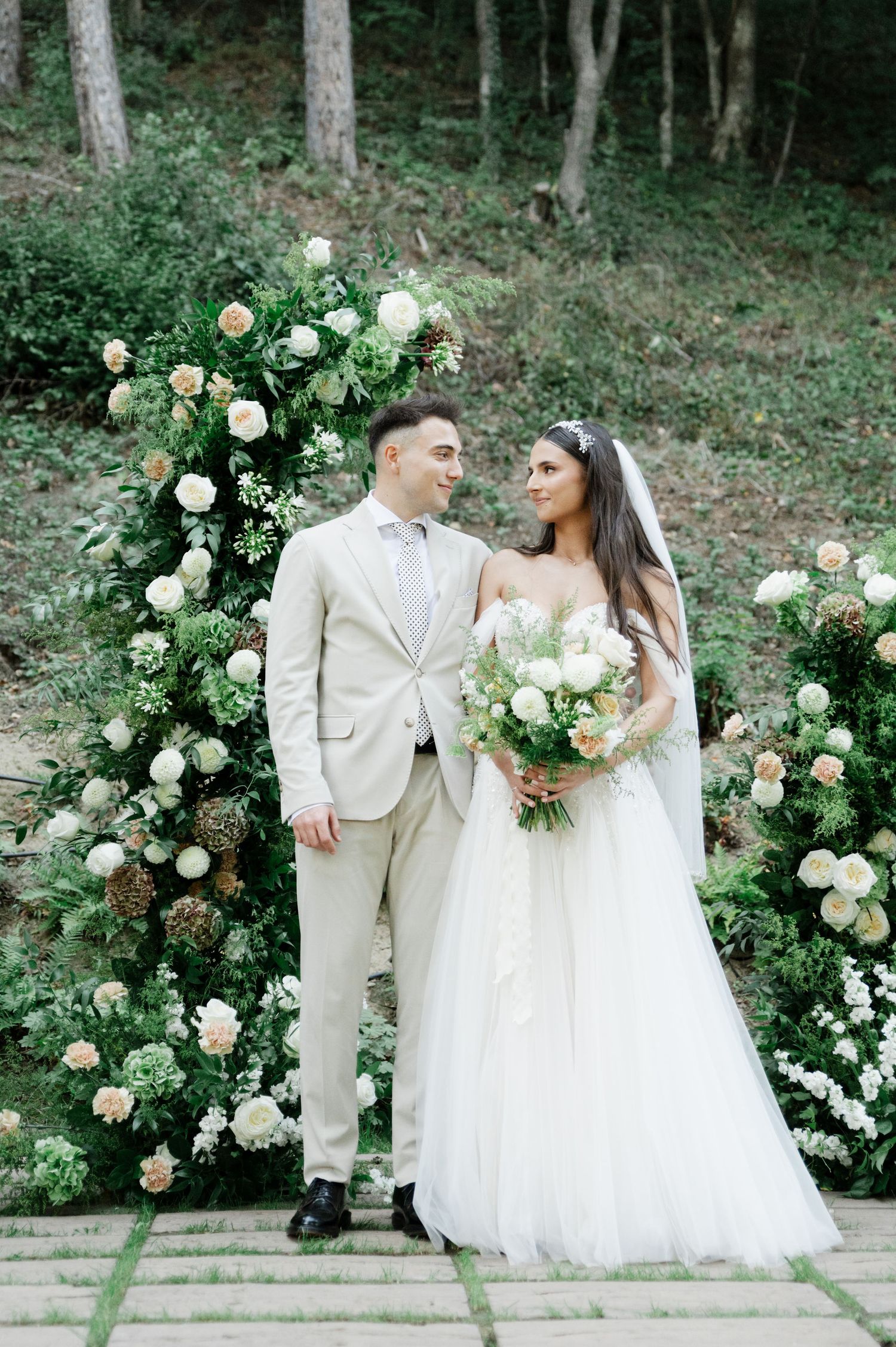 Romantic wedding portrait against a floral arch with cream roses in a forest setting.