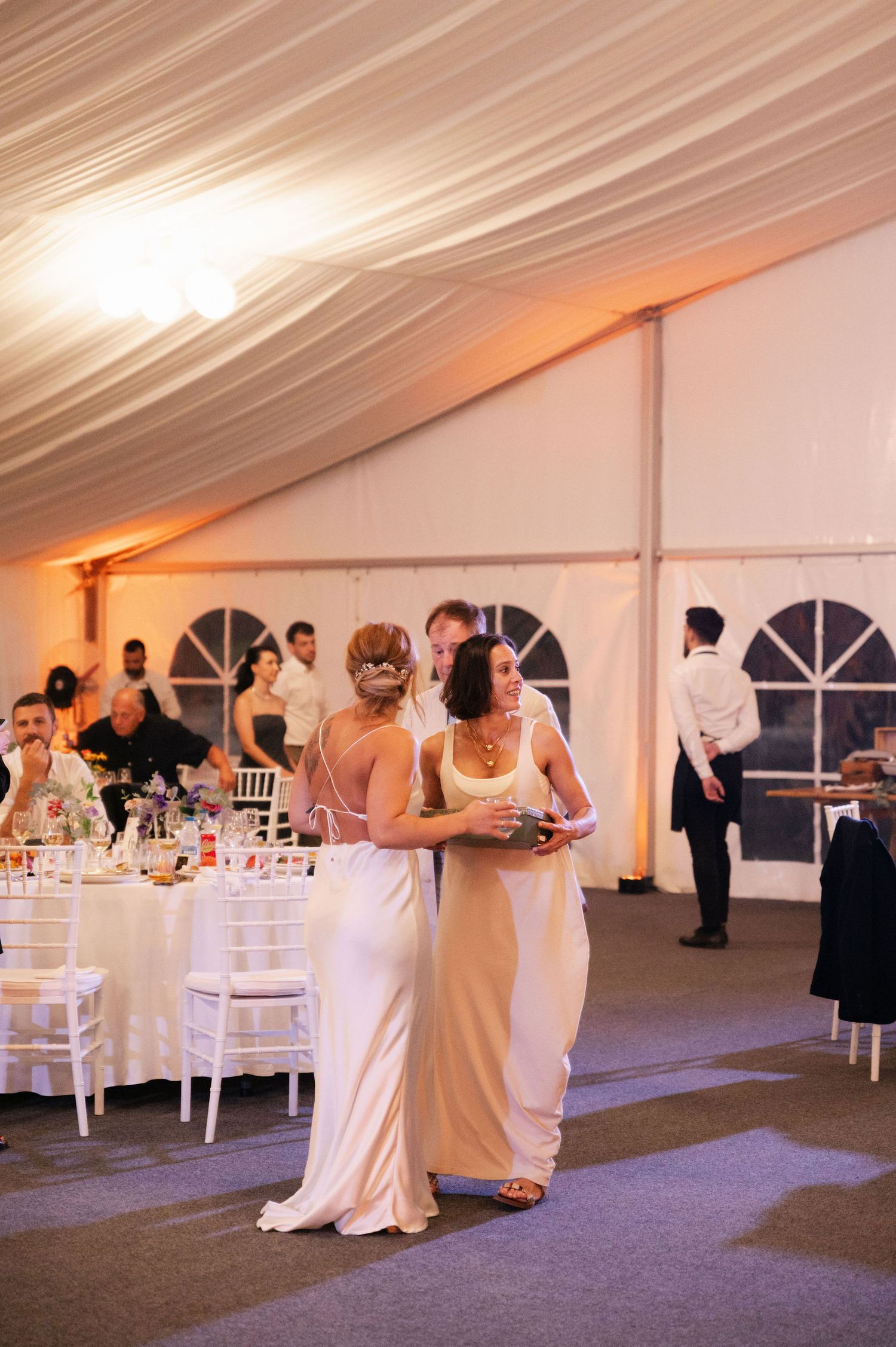 Two guests in elegant dresses share a moment during an evening wedding reception under a draped tent ceiling.