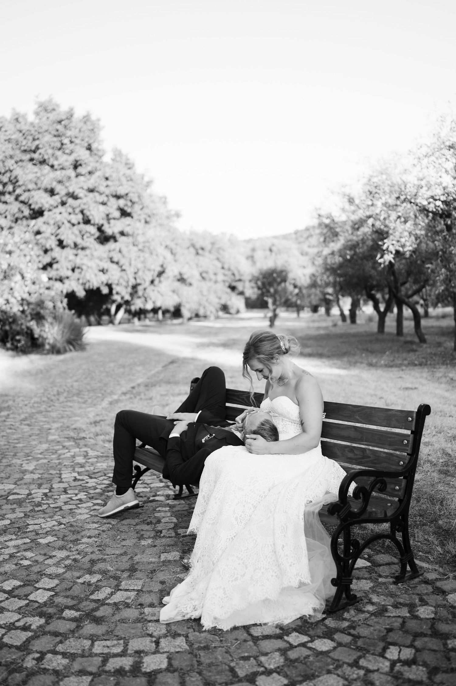 A romantic couple in formal attire share an intimate moment on a park bench surrounded by trees.