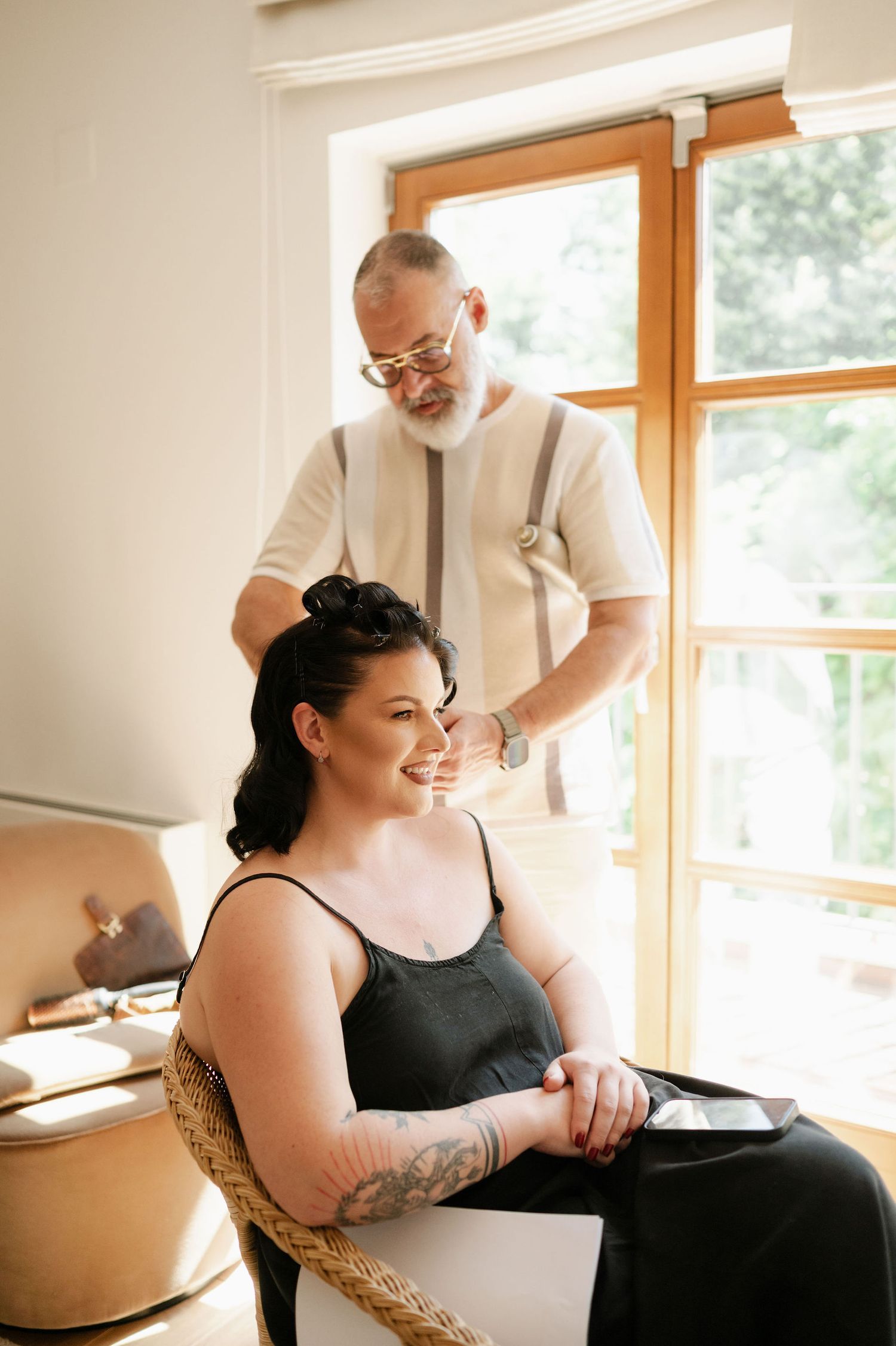 Makeup artist working on client's hair while seated in natural light from window.