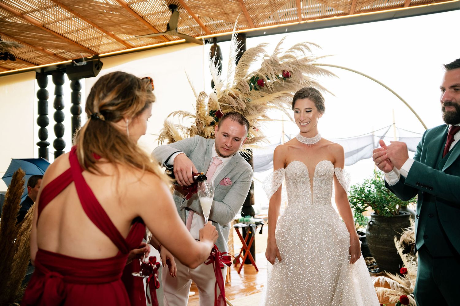 Wedding party gathers in a sunlit venue decorated with dried pampas grass and wooden architectural details.
