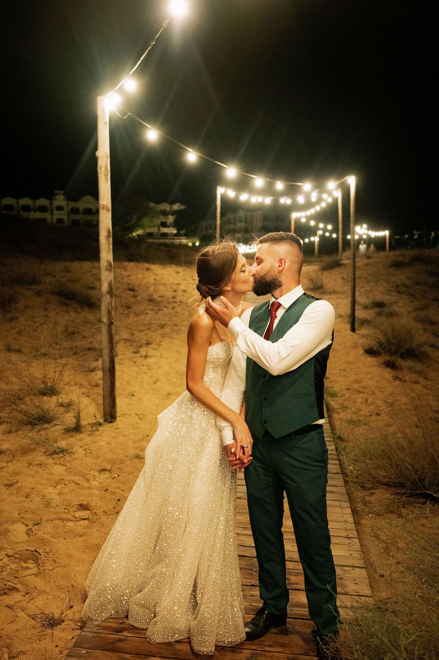A tender wedding moment captured under string lights on a beachside boardwalk.