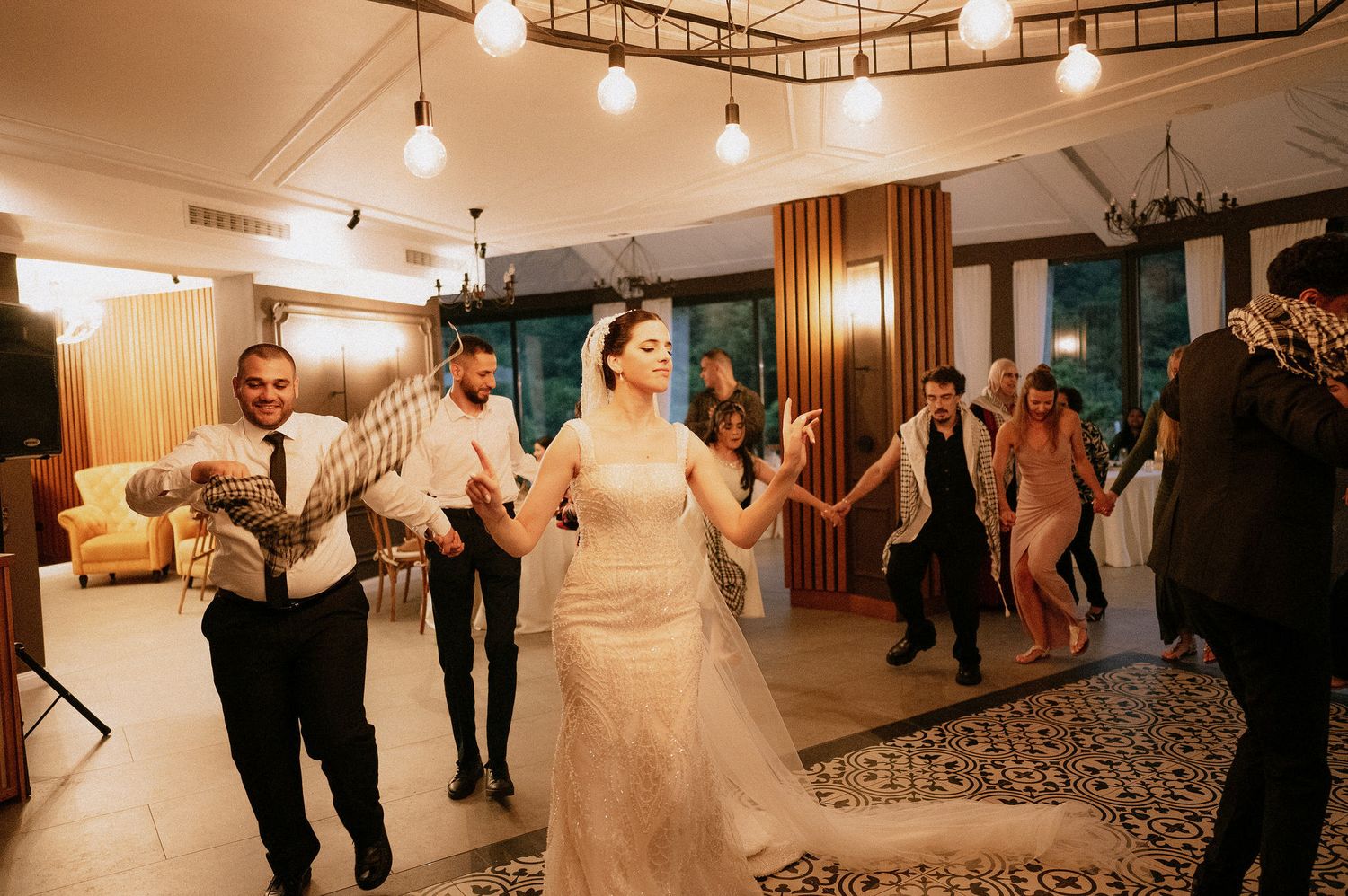 Panoramic view of wedding guests dancing in an indoor venue with decorative lighting.