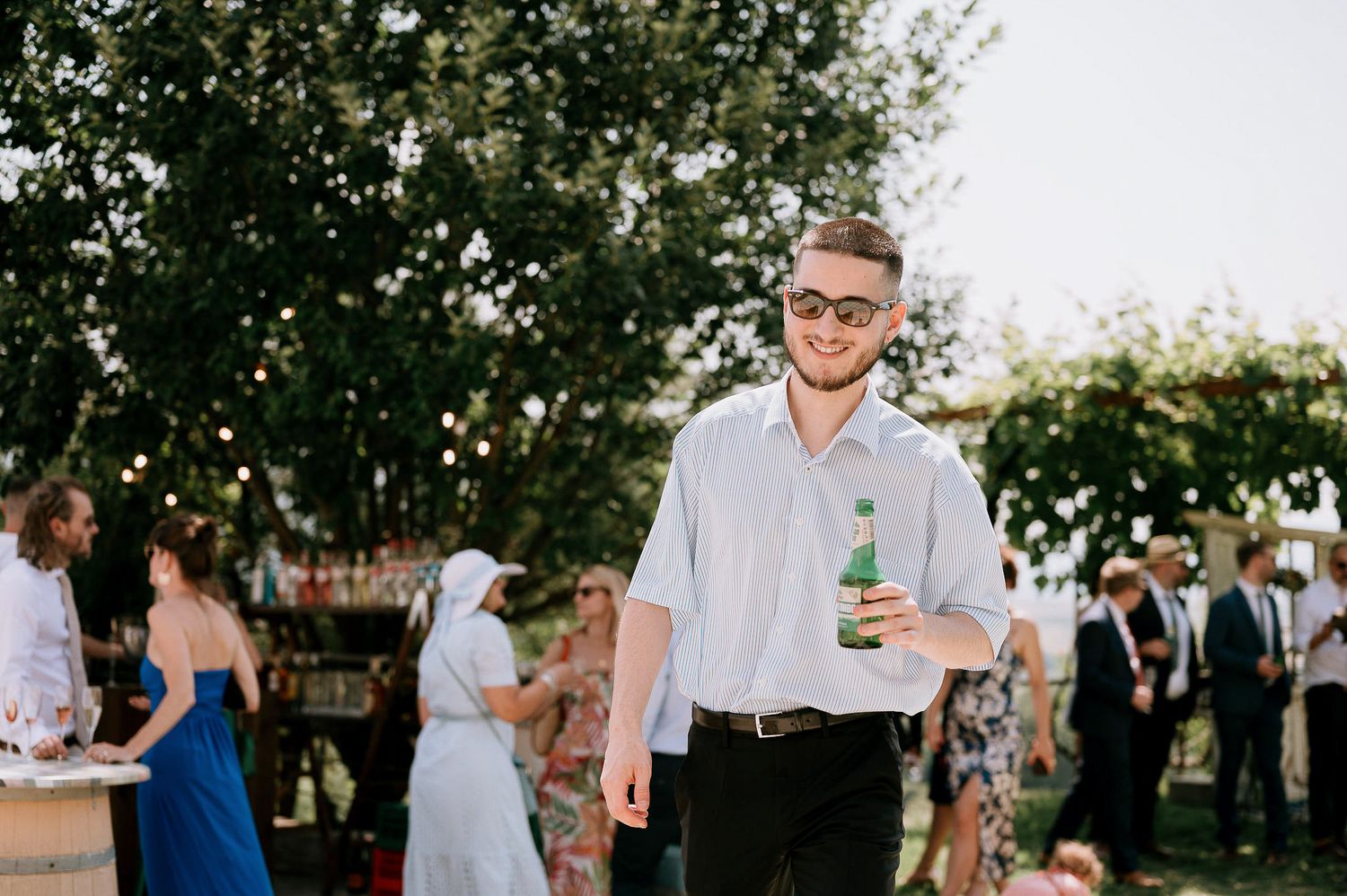 A guest at an outdoor wedding reception holds a drink while mingling with others.