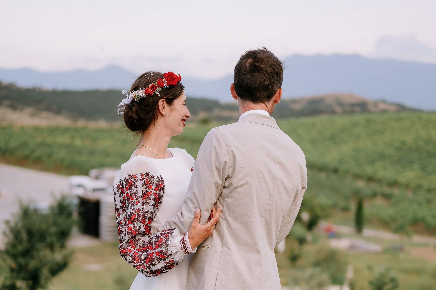 Wedding couple in white and beige stand together on grassy hillside with mountain landscape behind them.