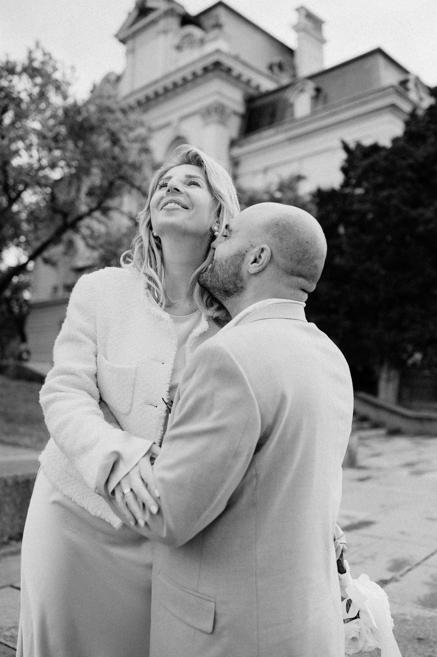 A couple in matching white attire shares an affectionate moment near a historic structure.