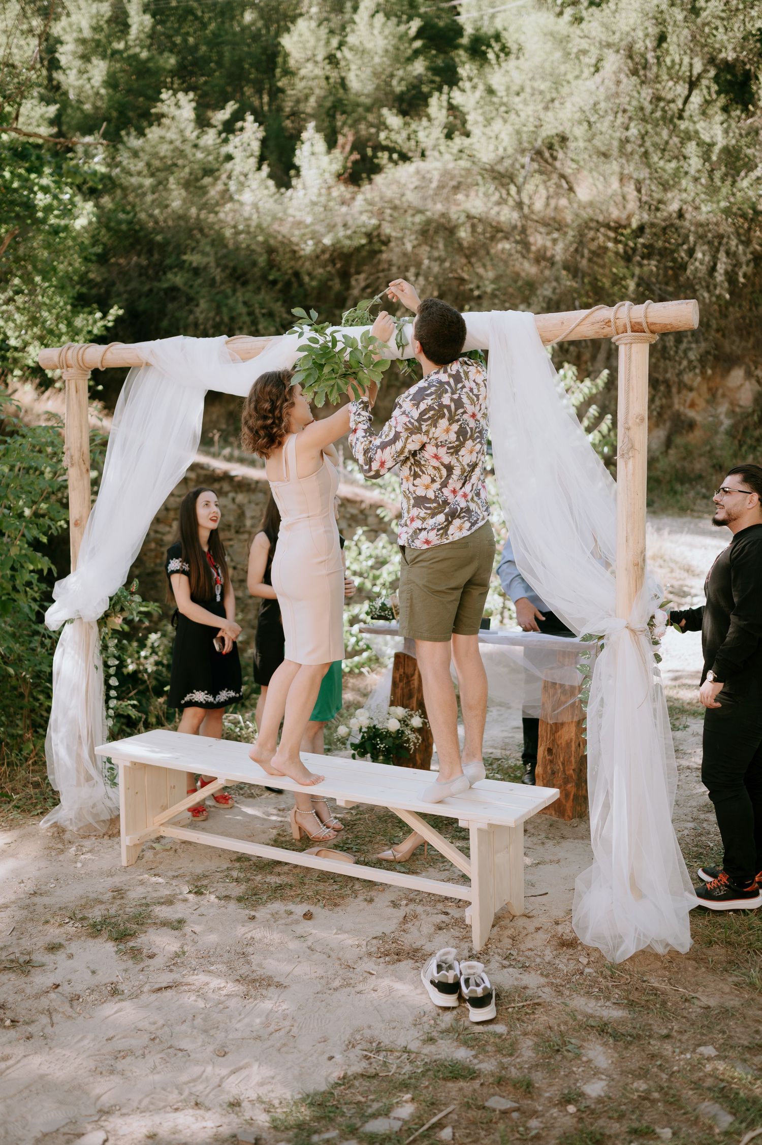 People setting up white fabric decorations on wooden posts for an outdoor ceremony.