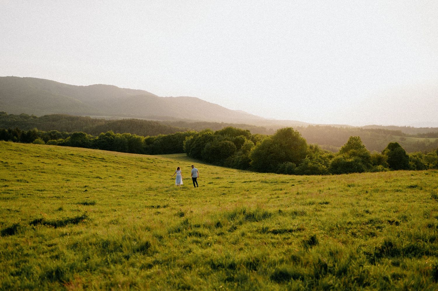 Two figures strolling together in a vast green field with mountains in background.