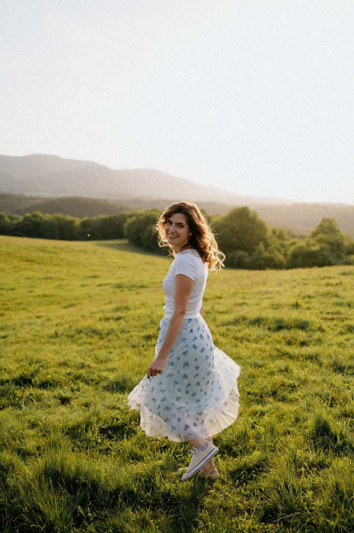 A flowing white dress twirls gracefully in a sunlit meadow with rolling hills in the background.