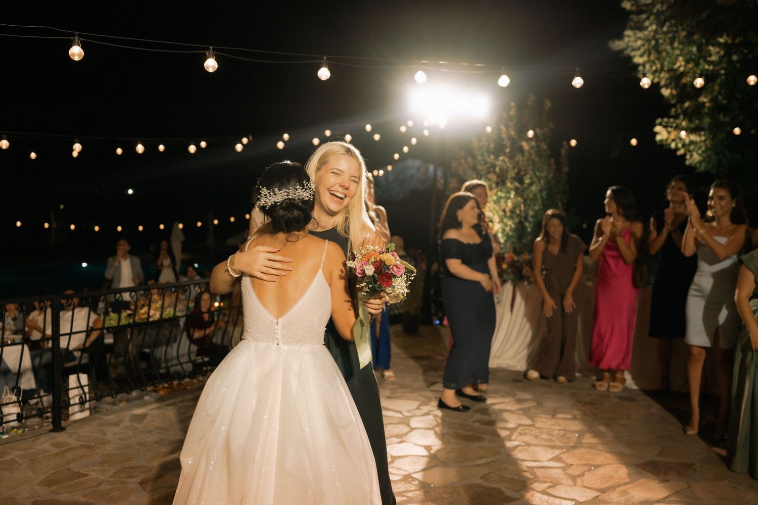 String lights illuminate an outdoor evening wedding reception with guests mingling and dancing on a stone patio.
