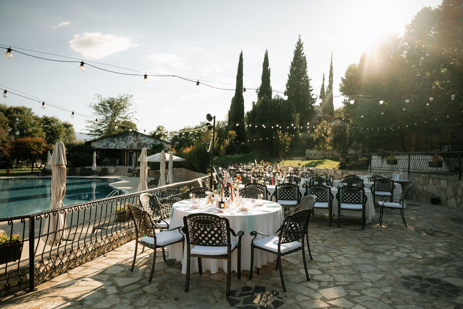String lights illuminate an outdoor patio dining area with tables and chairs at sunset.