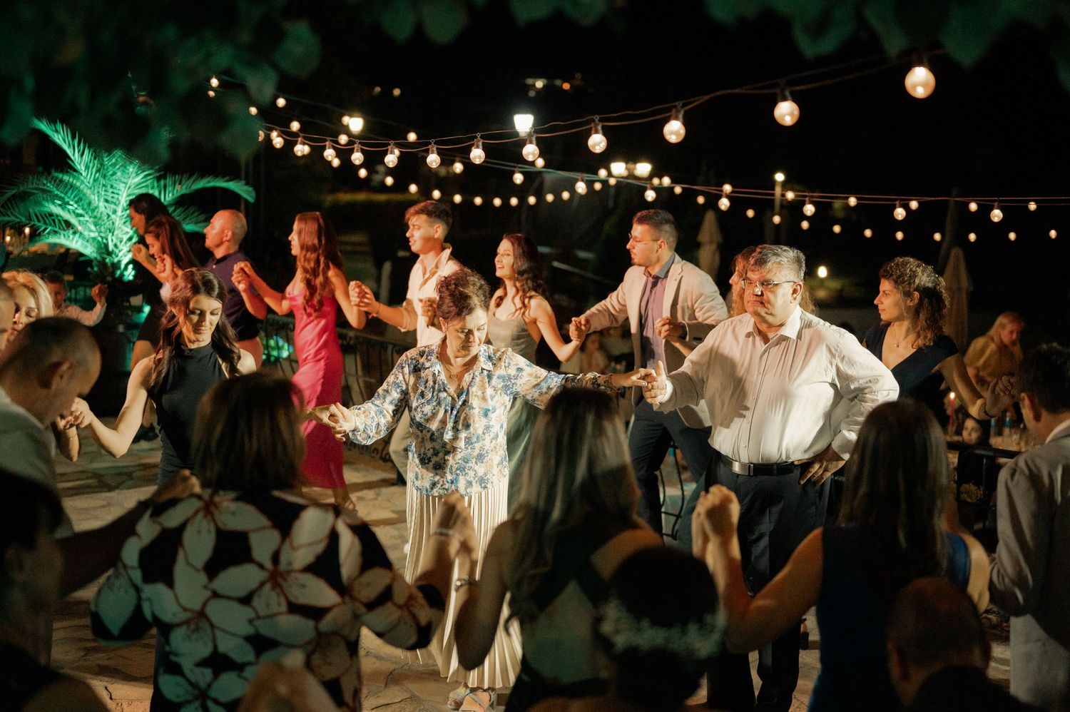 Wedding guests dance together under twinkling string lights during an evening outdoor reception celebration.