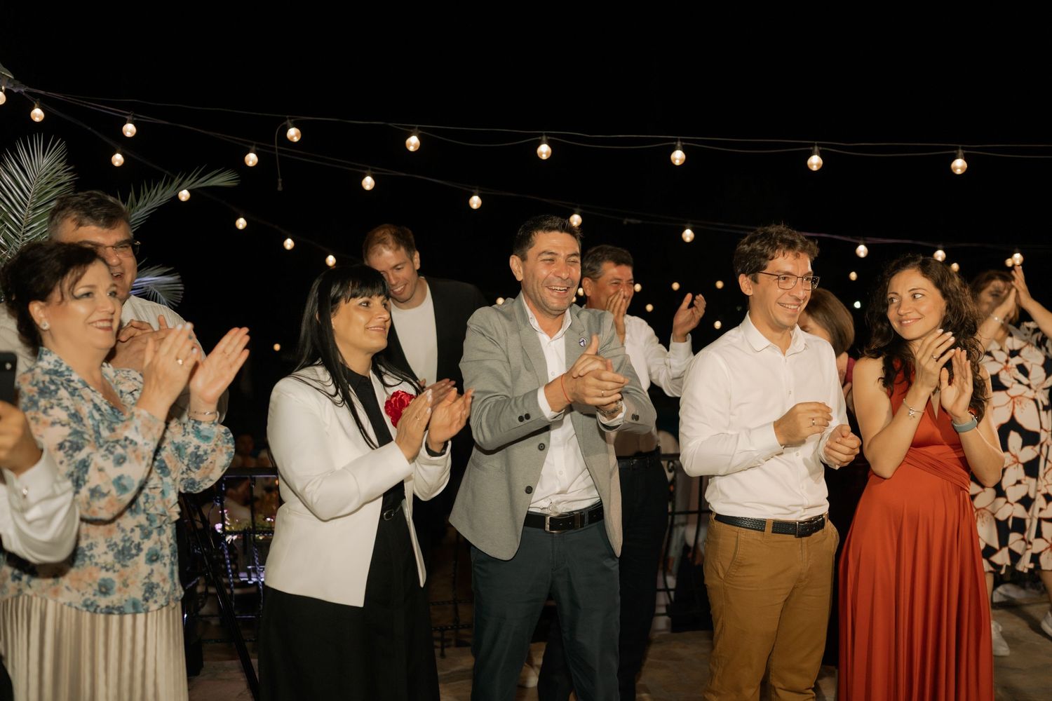 A group of wedding guests dance and celebrate at a nighttime outdoor reception with string lights overhead.