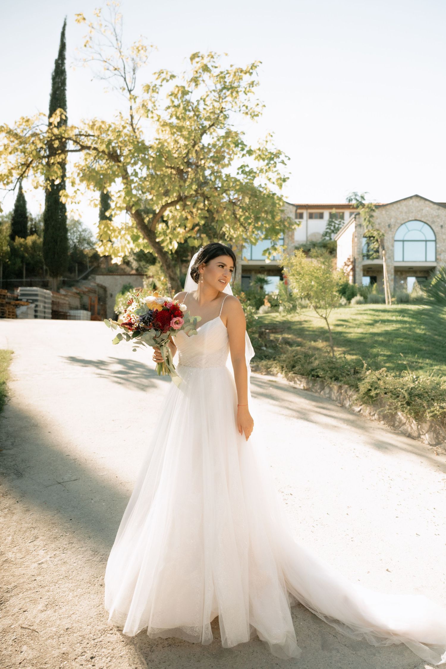 Bride in flowing white tulle wedding gown twirls gracefully on a sunlit pathway surrounded by autumn trees.