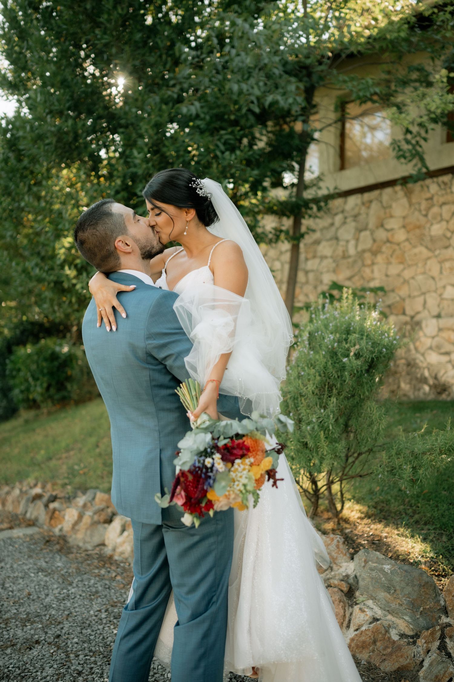 Newlywed couple shares romantic moment in garden setting with colorful bouquet and stone wall backdrop at sunset.