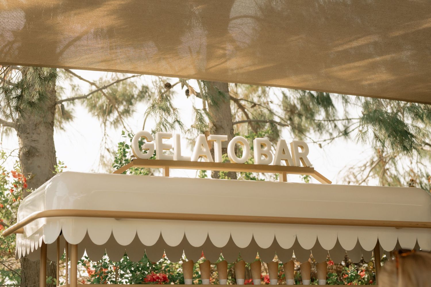 Elegant outdoor gelato bar with gold signage under a shaded canopy surrounded by lush greenery.