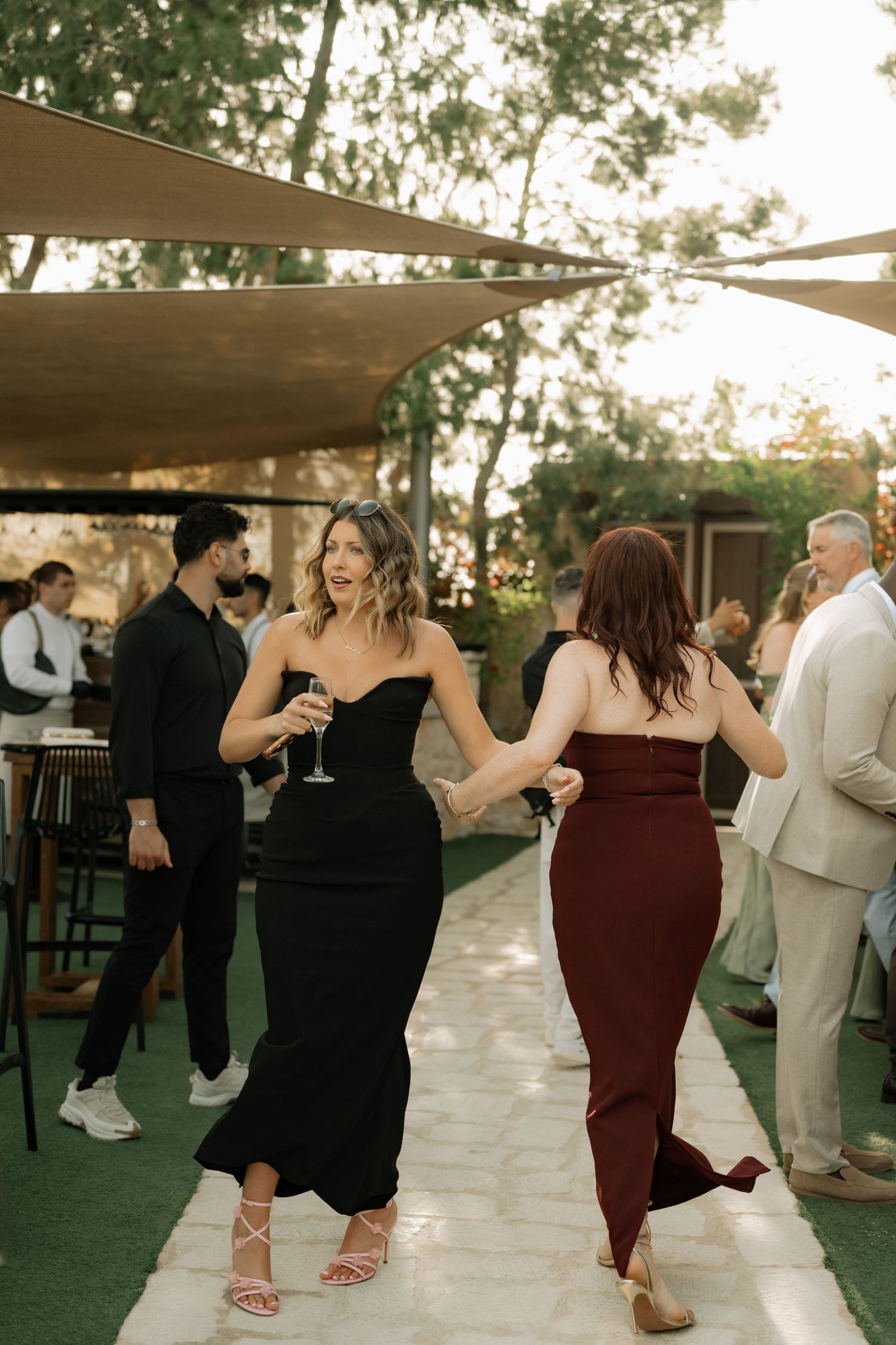 Two women in elegant black and burgundy gowns dancing at an outdoor wedding reception at golden hour.