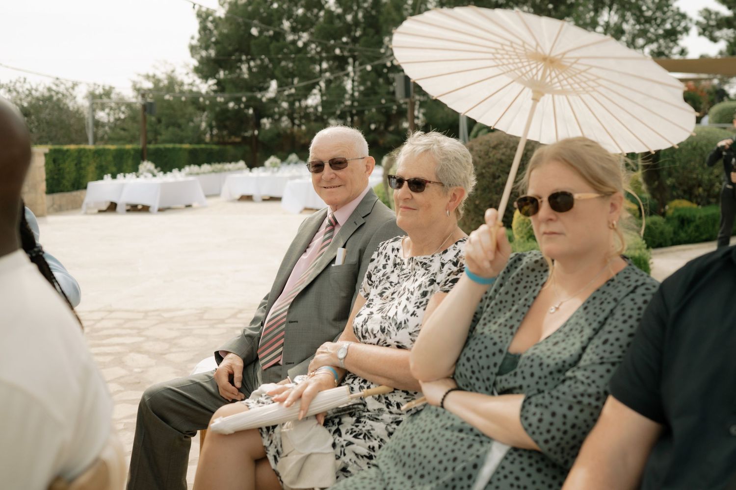 Wedding guests seated outdoors, including a woman holding a white parasol umbrella, enjoying an elegant garden ceremony.