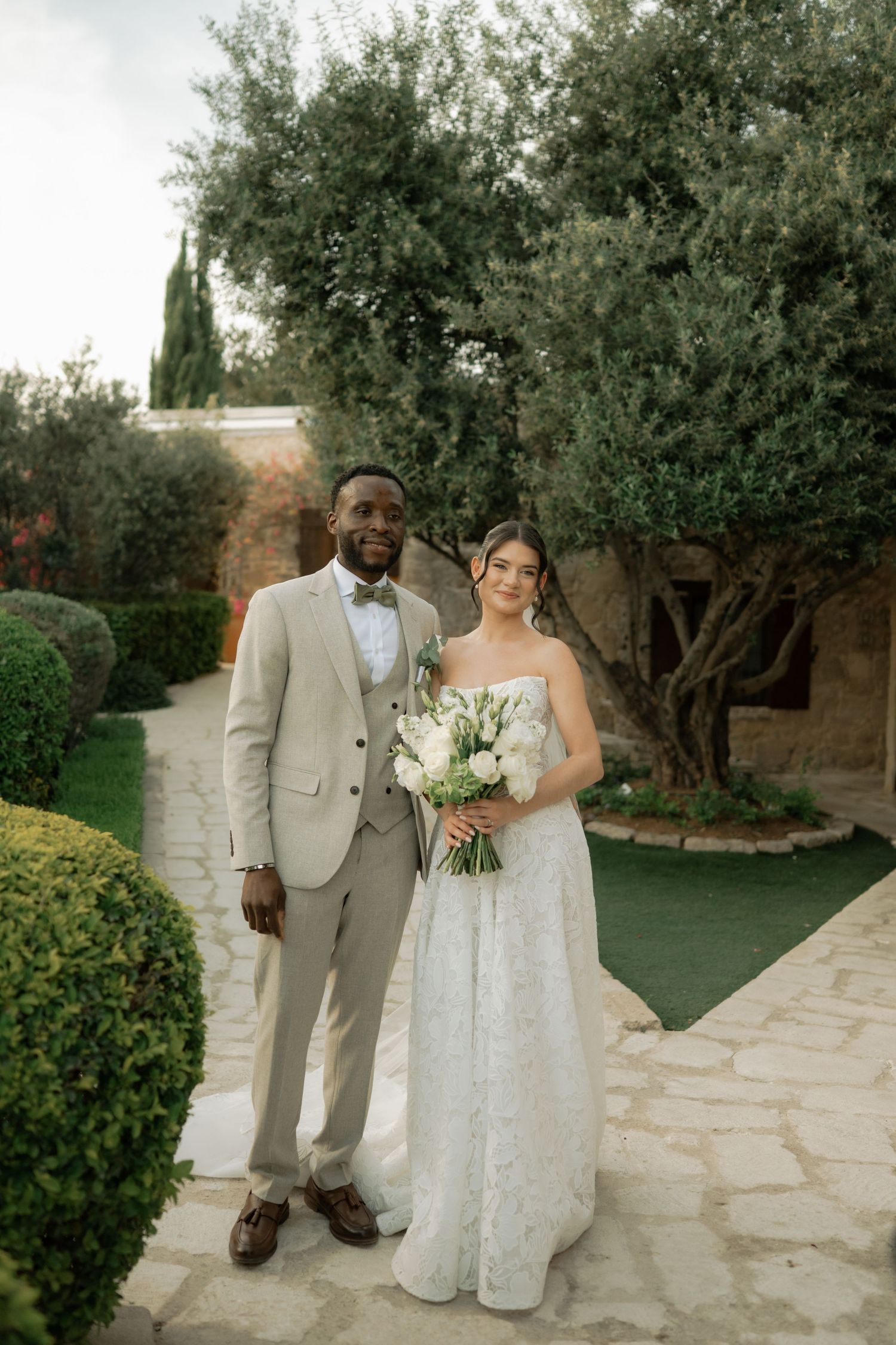 Interracial couple posing in garden on wedding day, groom in beige suit, bride in white lace gown holding bouquet.