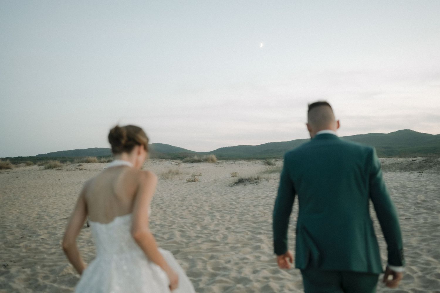 Bride and groom walking hand in hand on sandy beach dunes at dusk during romantic wedding photoshoot.
