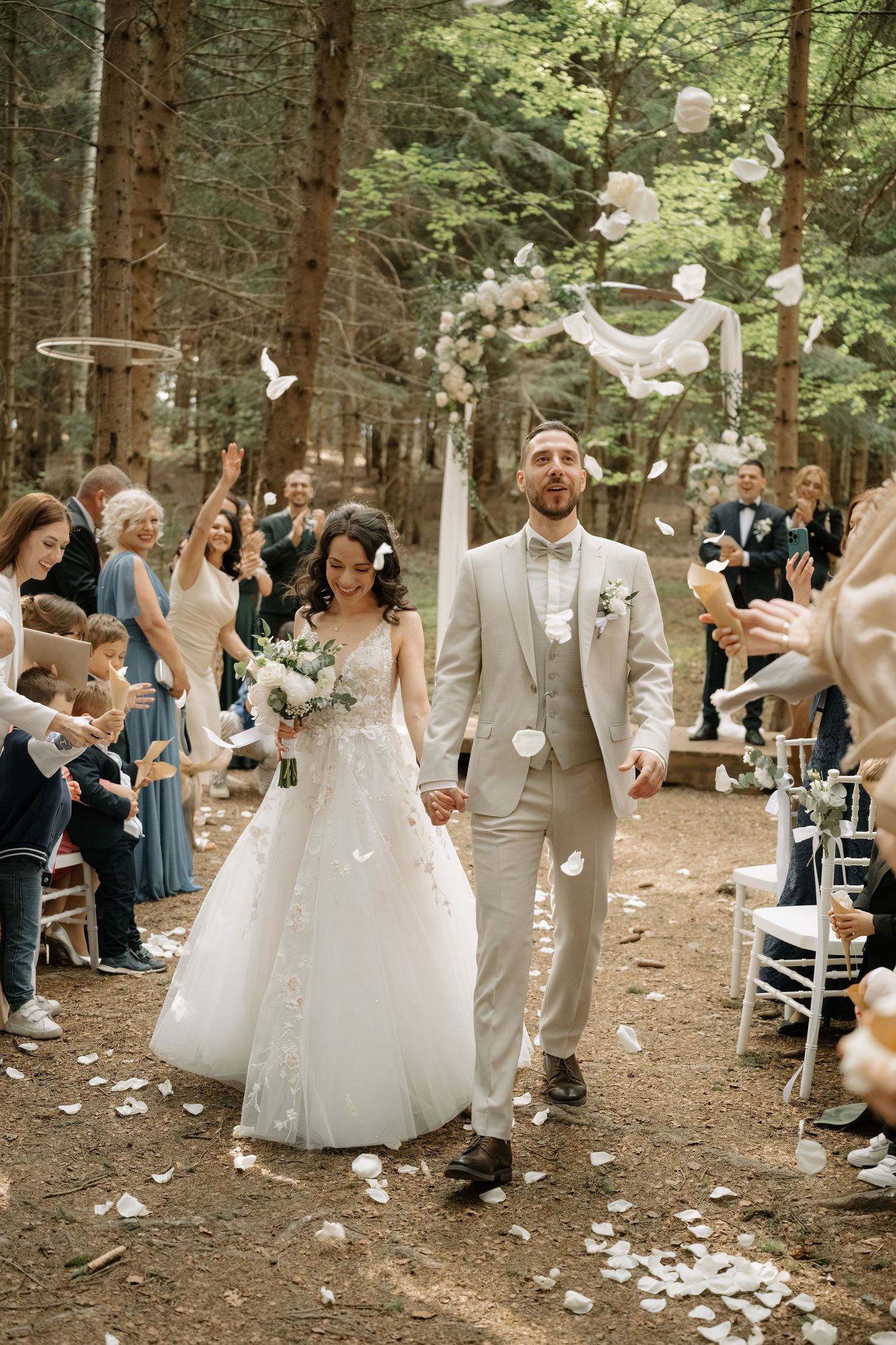 Bride and groom walk down forest aisle as guests toss white petals at outdoor woodland wedding ceremony.