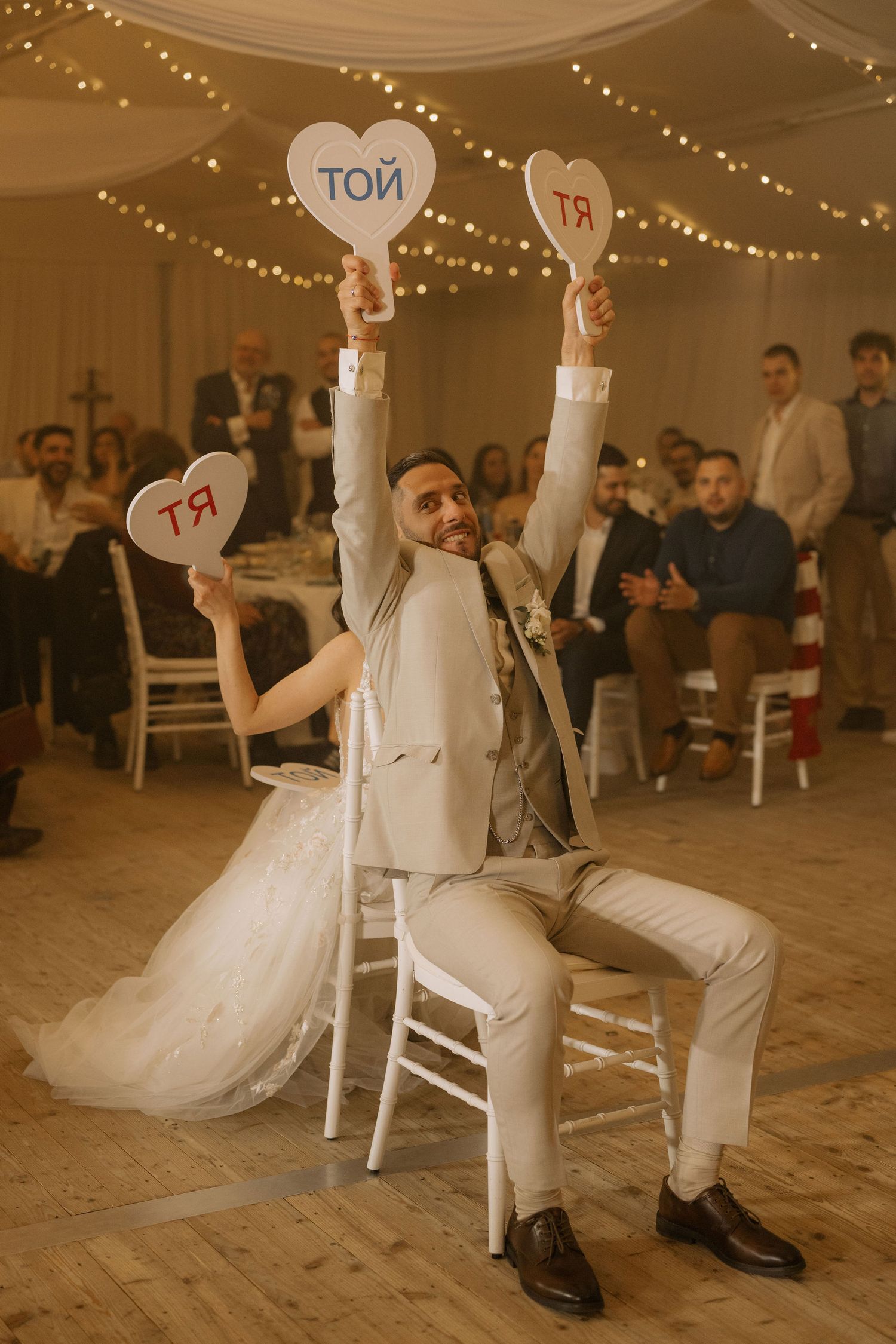 Bride and groom hold heart-shaped signs reading TOY and YA at wedding reception with fairy lights and guests.