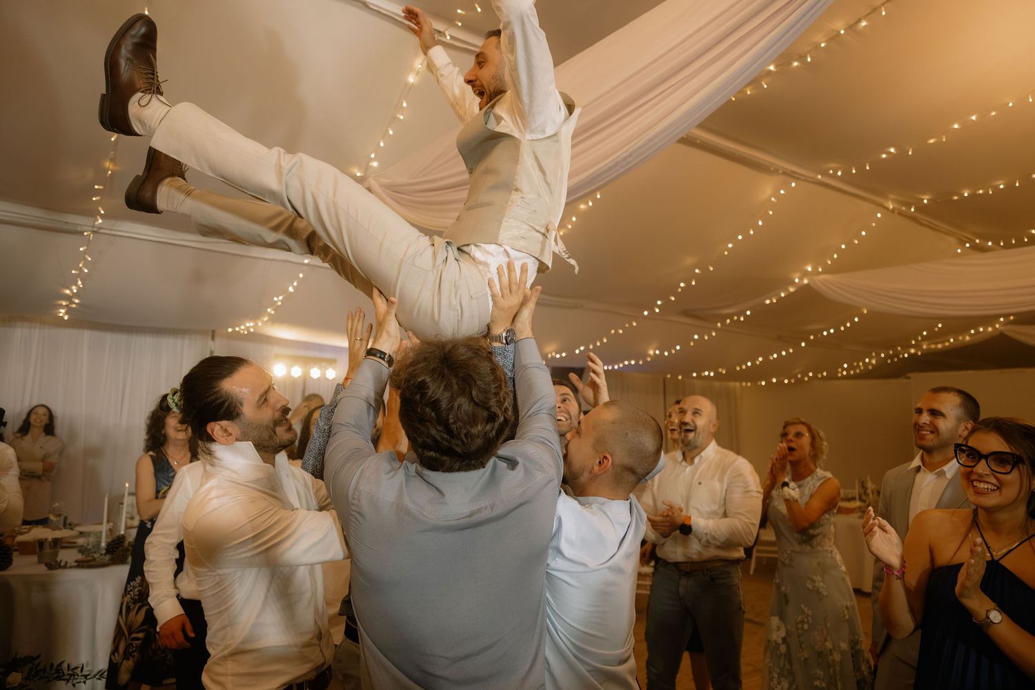 Guests lifting a man in the air during a lively wedding reception celebration indoors with fairy lights.