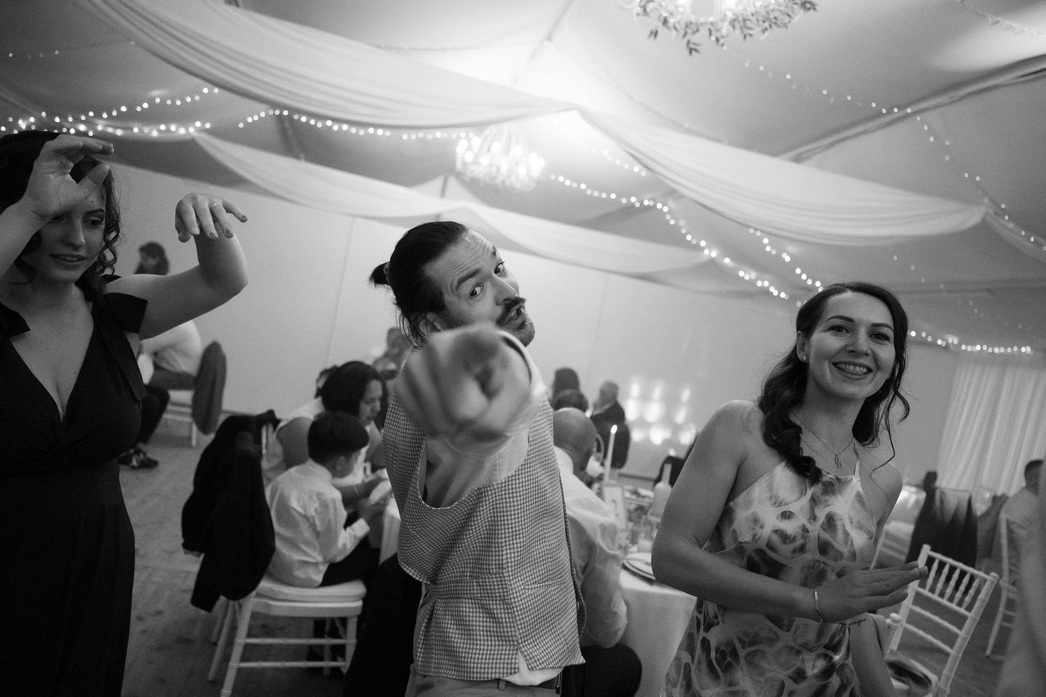 Black and white photo of guests dancing and laughing at a wedding reception in a decorated marquee tent.