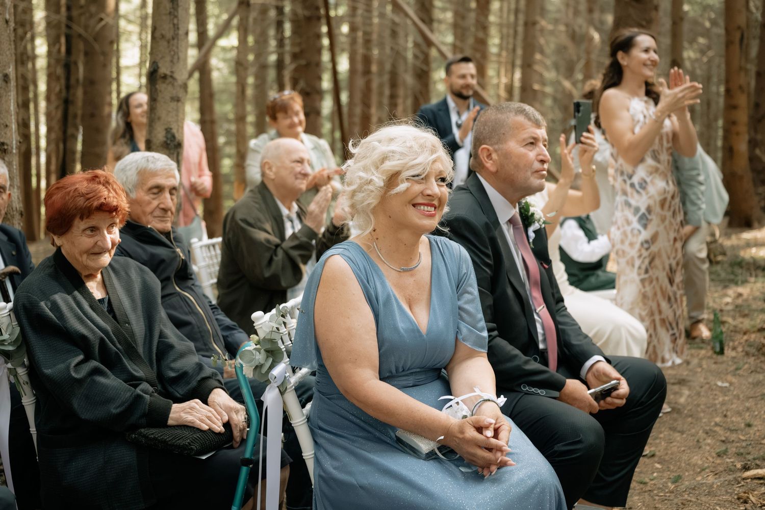 Wedding guests smiling and laughing during an outdoor forest ceremony, seated on chairs among tall pine trees.