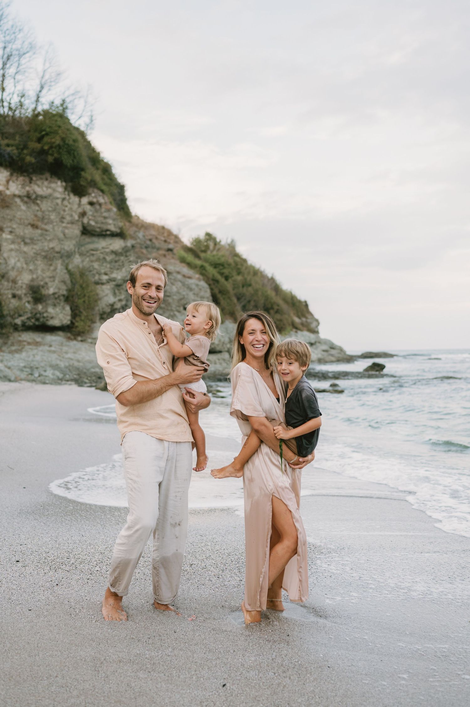 Happy family of four posing on a sandy beach with rocky cliffs, waves, and an overcast sky.