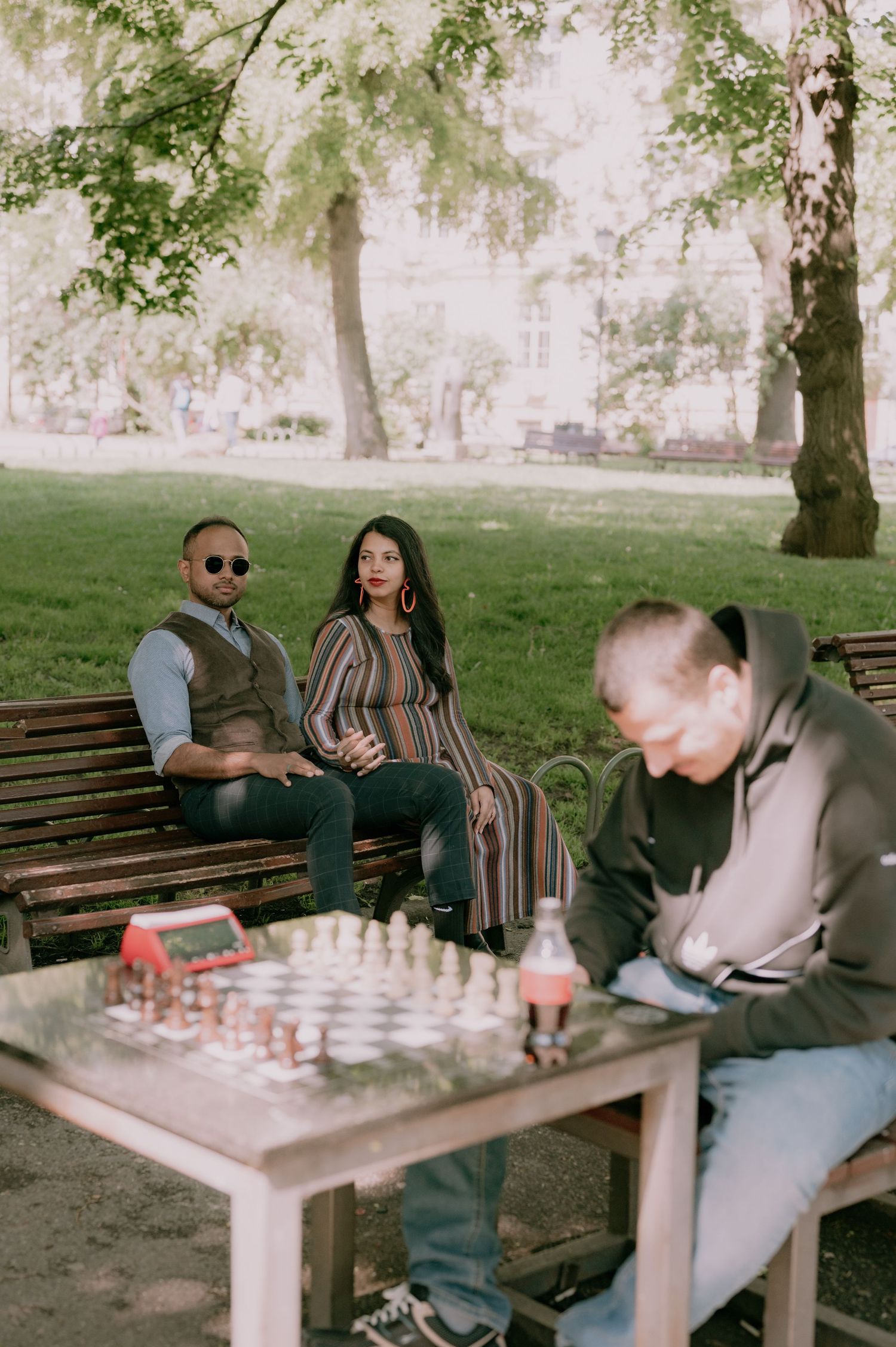 Two people sit on a park bench watching a man play chess on an outdoor stone table surrounded by lush green trees.