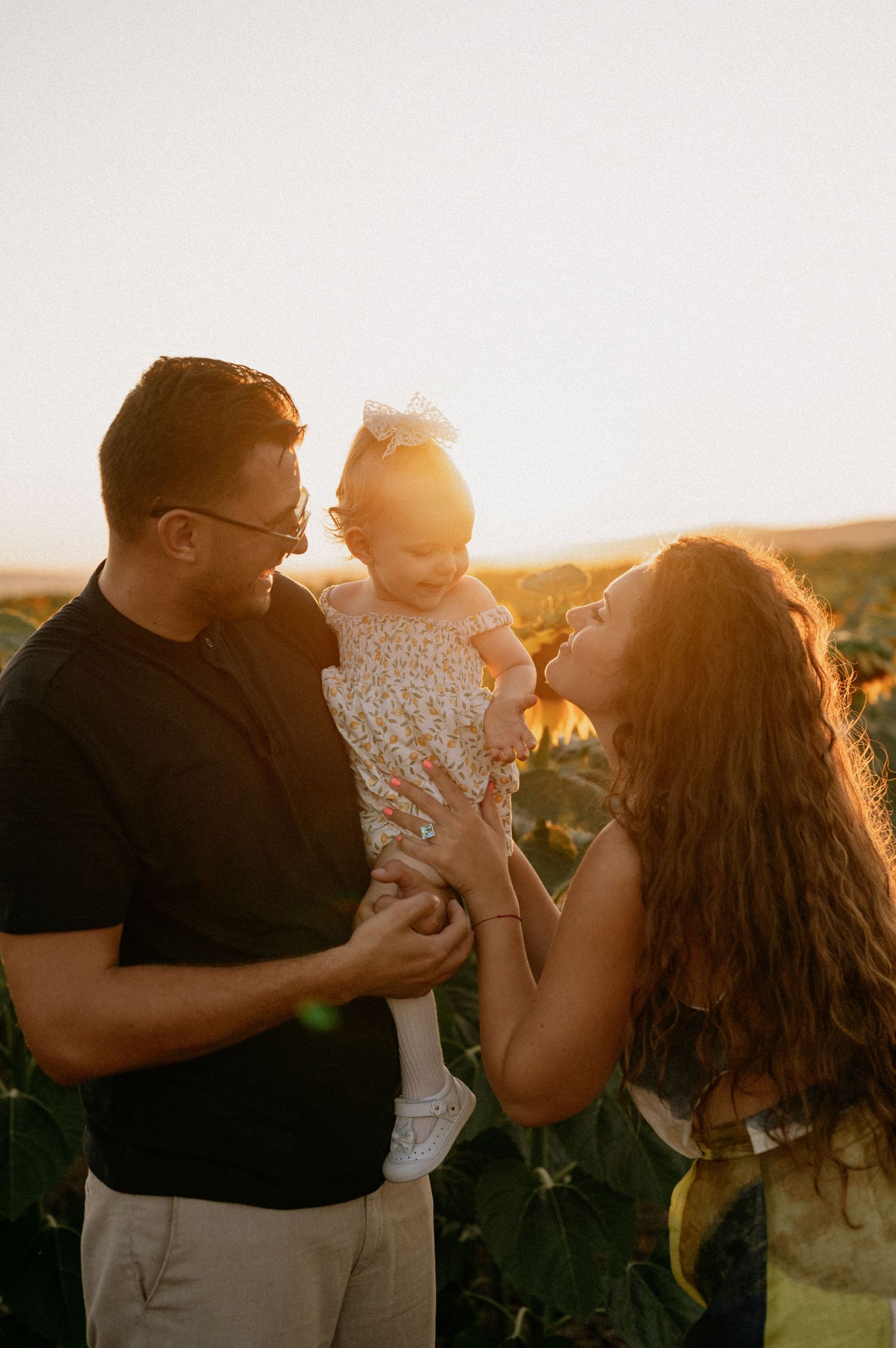 A couple smiles at their baby girl in a sunlit sunflower field during a golden hour family photo session.