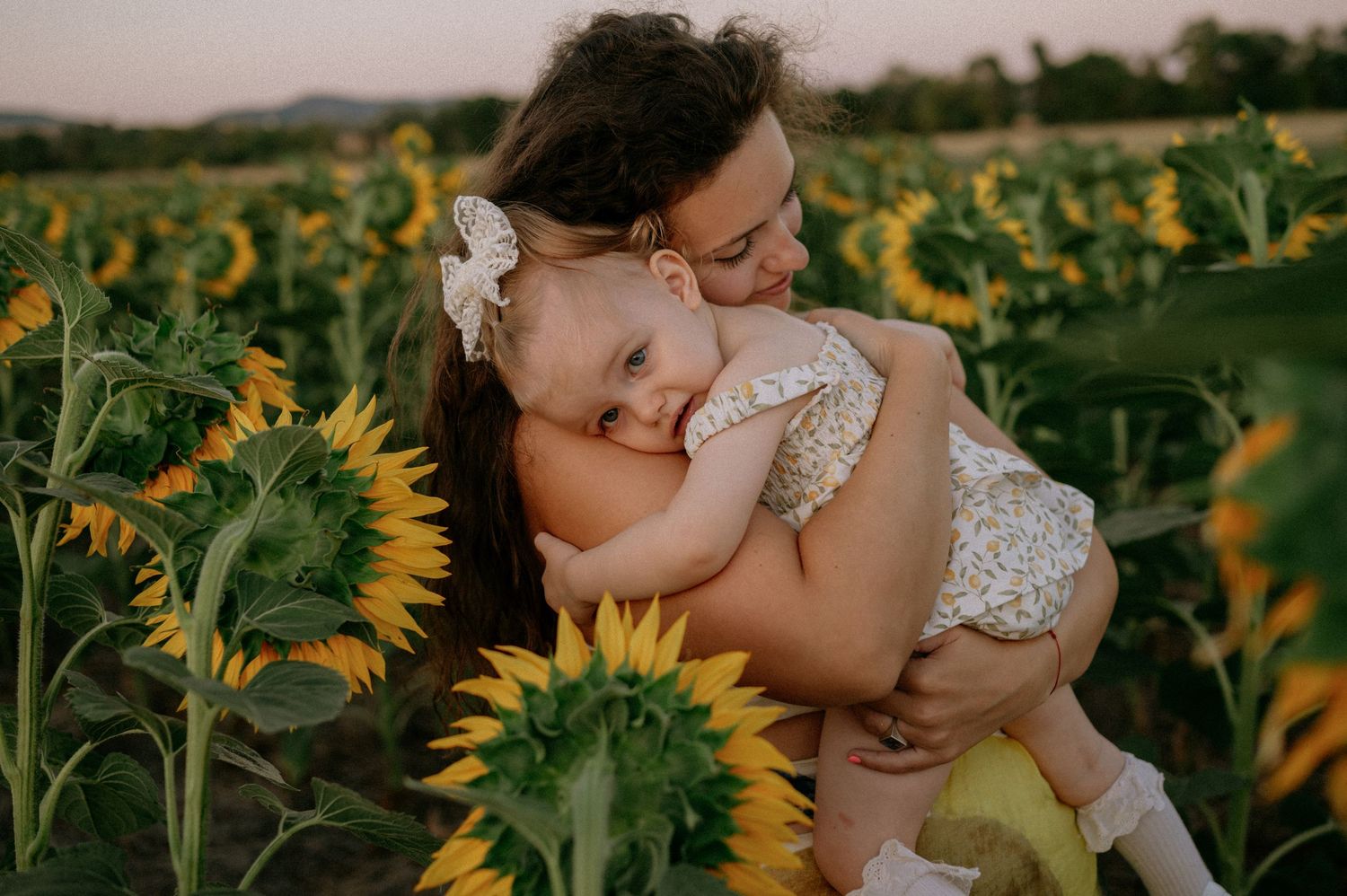 A mother lovingly holds her smiling baby in a vibrant sunflower field during golden hour.