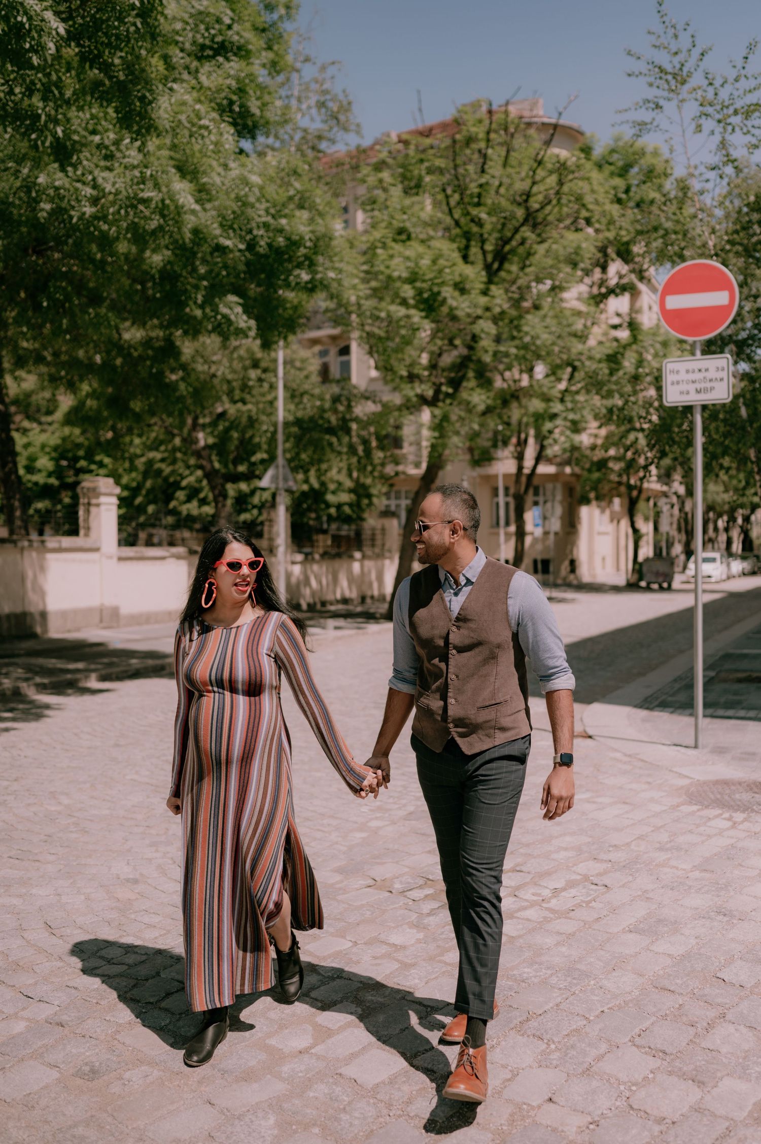 Pregnant woman in striped dress holds hands with man in vest walking on sunny urban street.