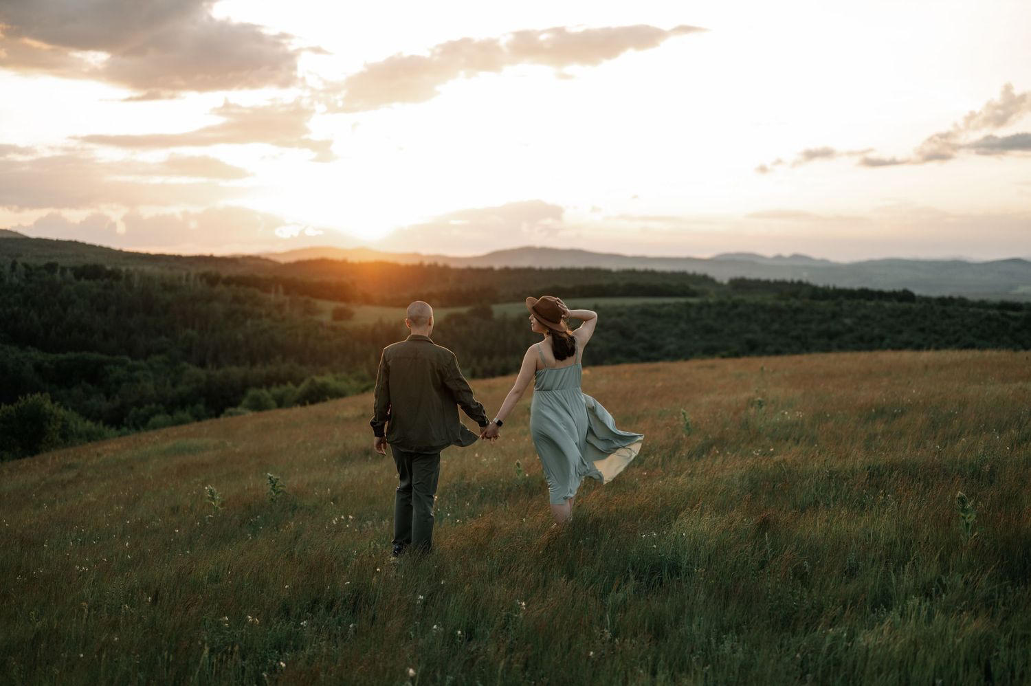 Couple holding hands walking through a lush green meadow toward a stunning golden sunset over rolling hills.