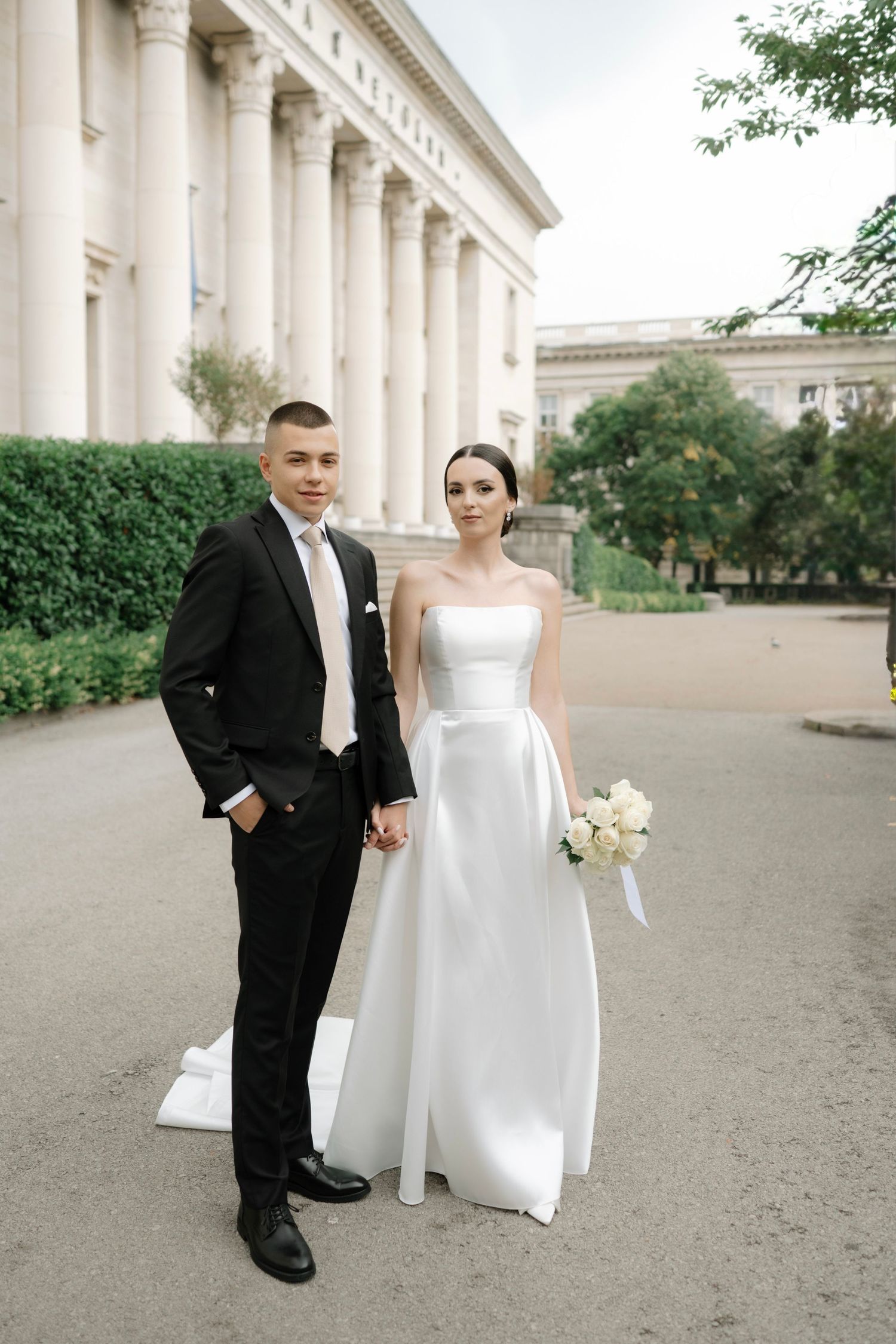 A wedding couple poses in front of a grand white columned building with classical architecture.
