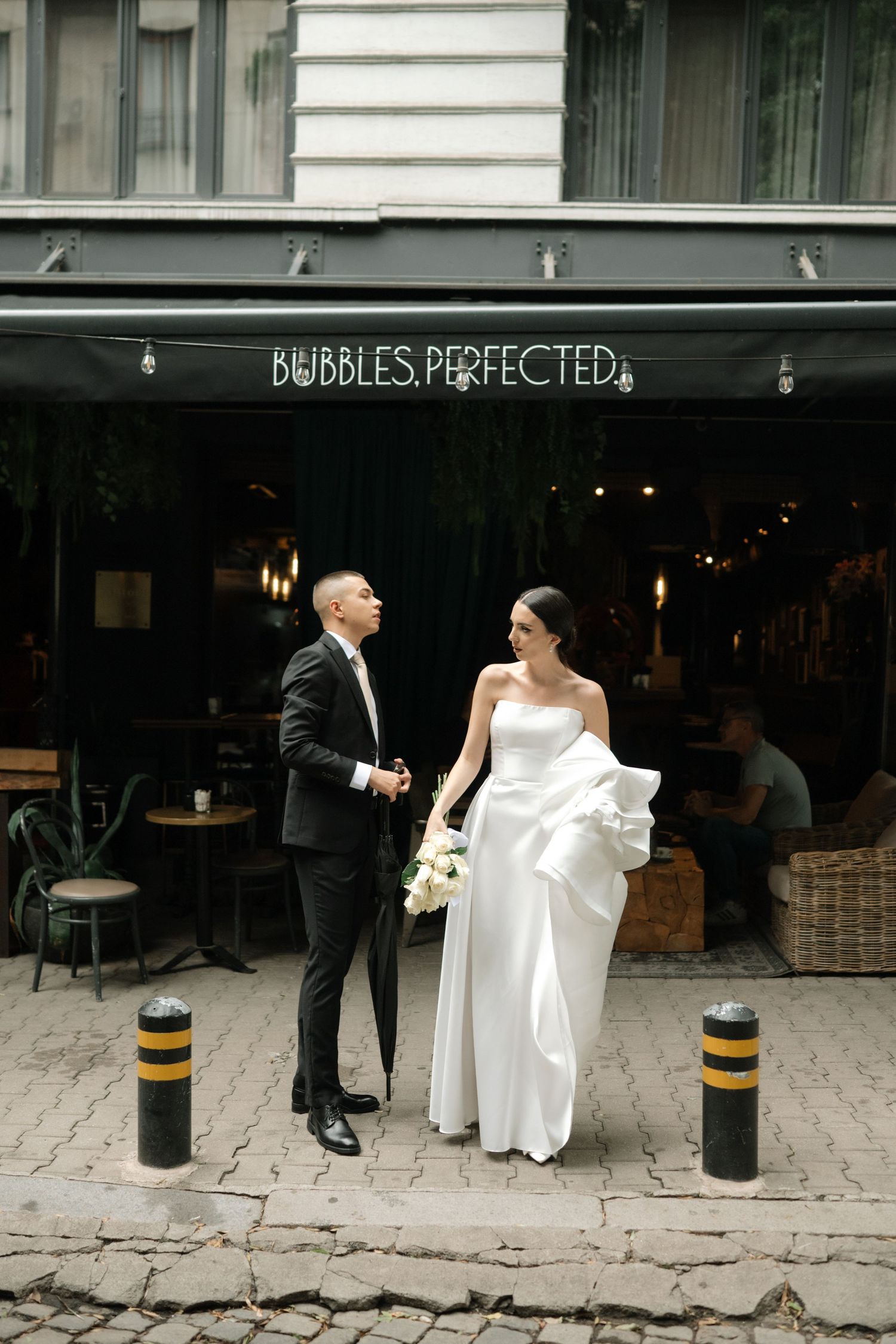 A couple in wedding attire stands outside a cafe called 'Bubbles Perfected' on a cobblestone street.