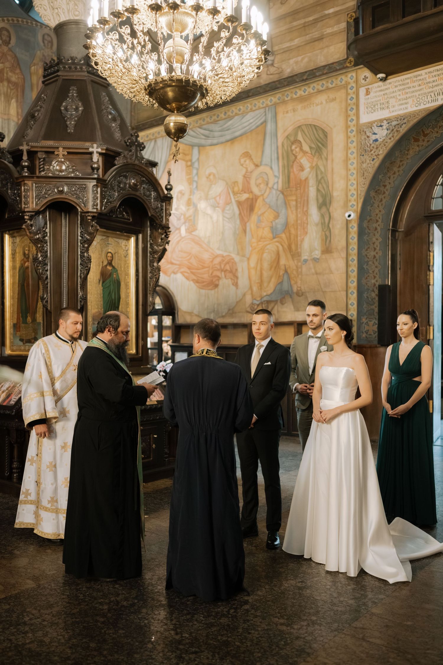 Traditional Orthodox wedding ceremony taking place in an ornate church with chandelier and religious artwork on walls.