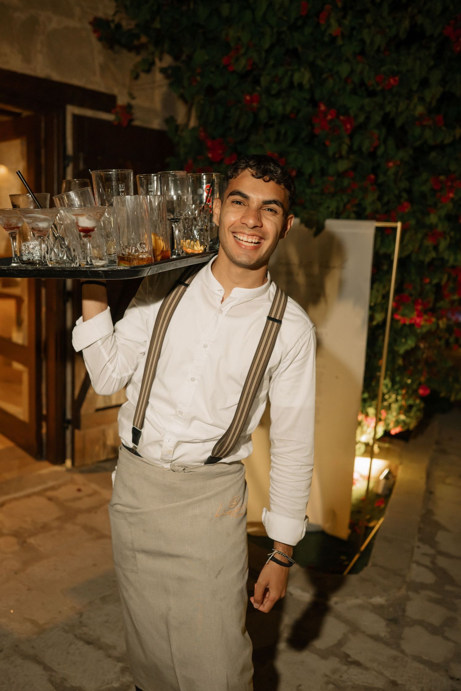 Smiling waiter in white shirt and suspenders carries a tray of glasses at an outdoor evening event.