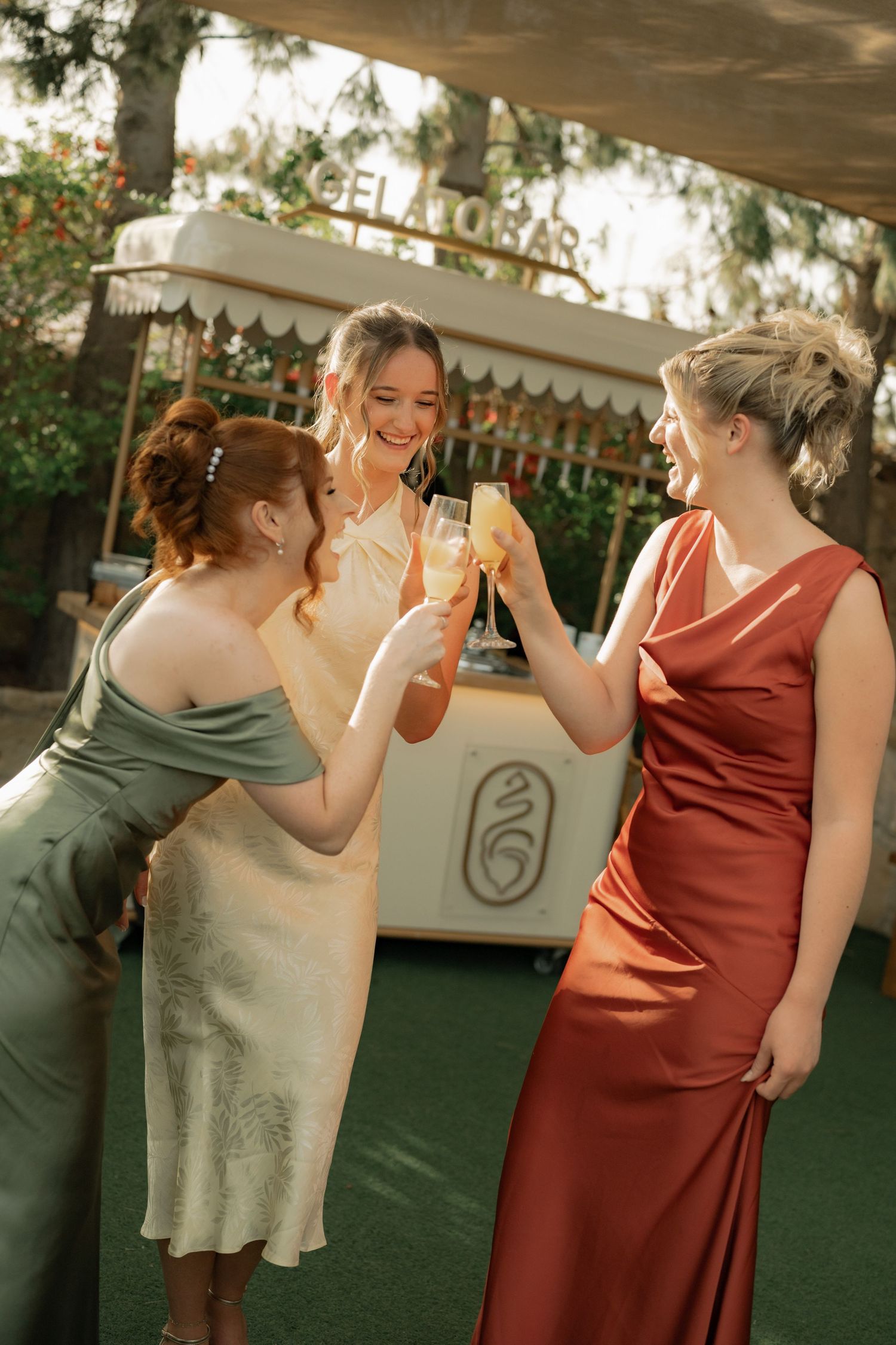 Three women in elegant dresses toasting with champagne glasses outdoors near a gelato bar.