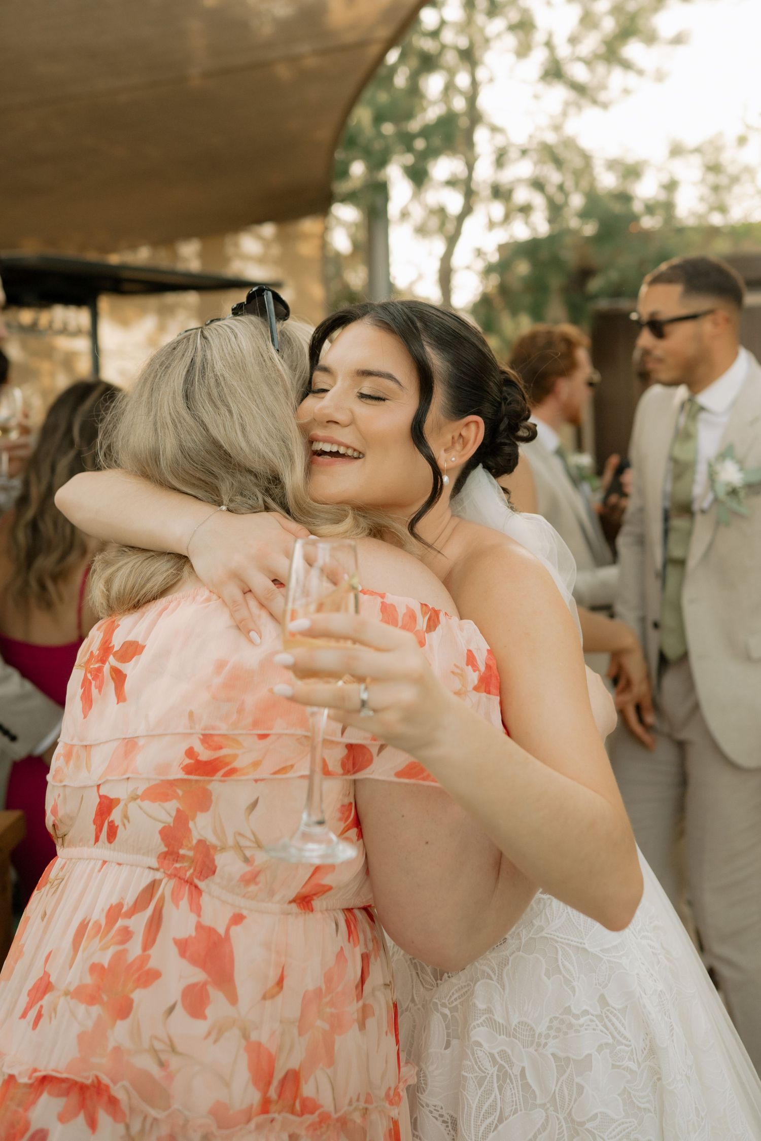 Bride in white lace gown hugs a guest in floral pink dress, laughing joyfully at an outdoor wedding reception.