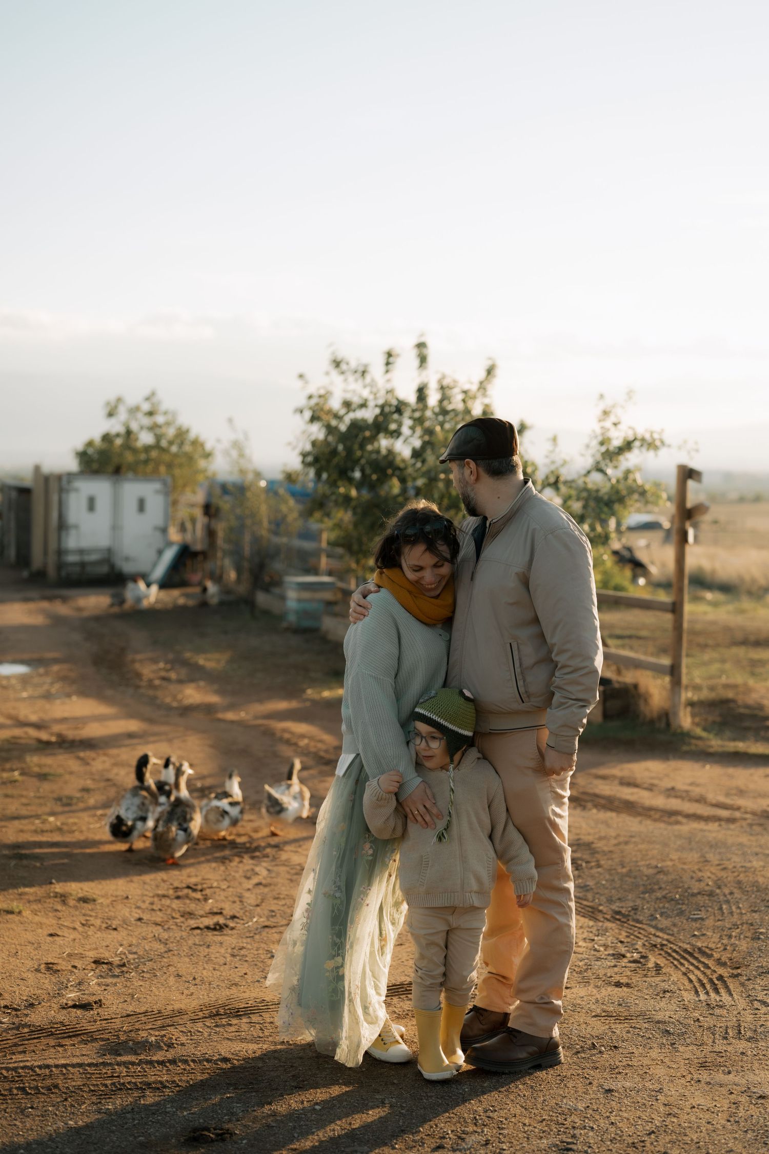 A couple in neutral toned outfits share a tender moment on dusty ground at sunset with trailers in the background.