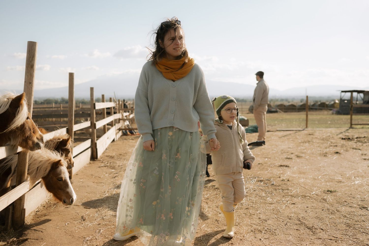 A person wearing a light blue dress stands in a desert landscape with wooden fences and structures in the background.