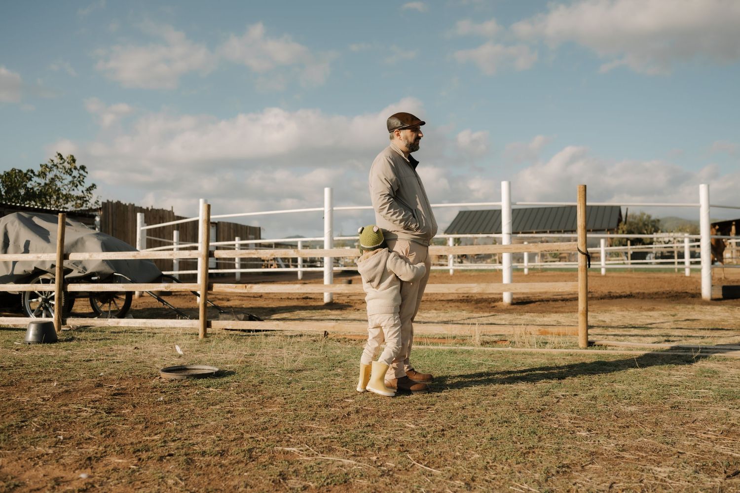 Someone stands in casual attire at a rustic ranch with white fencing and golden sunlight in the background.