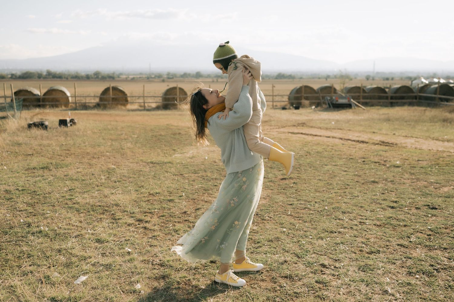 Person in a flowing white dress holds a bouquet while standing in a golden field with stone arches in the background.