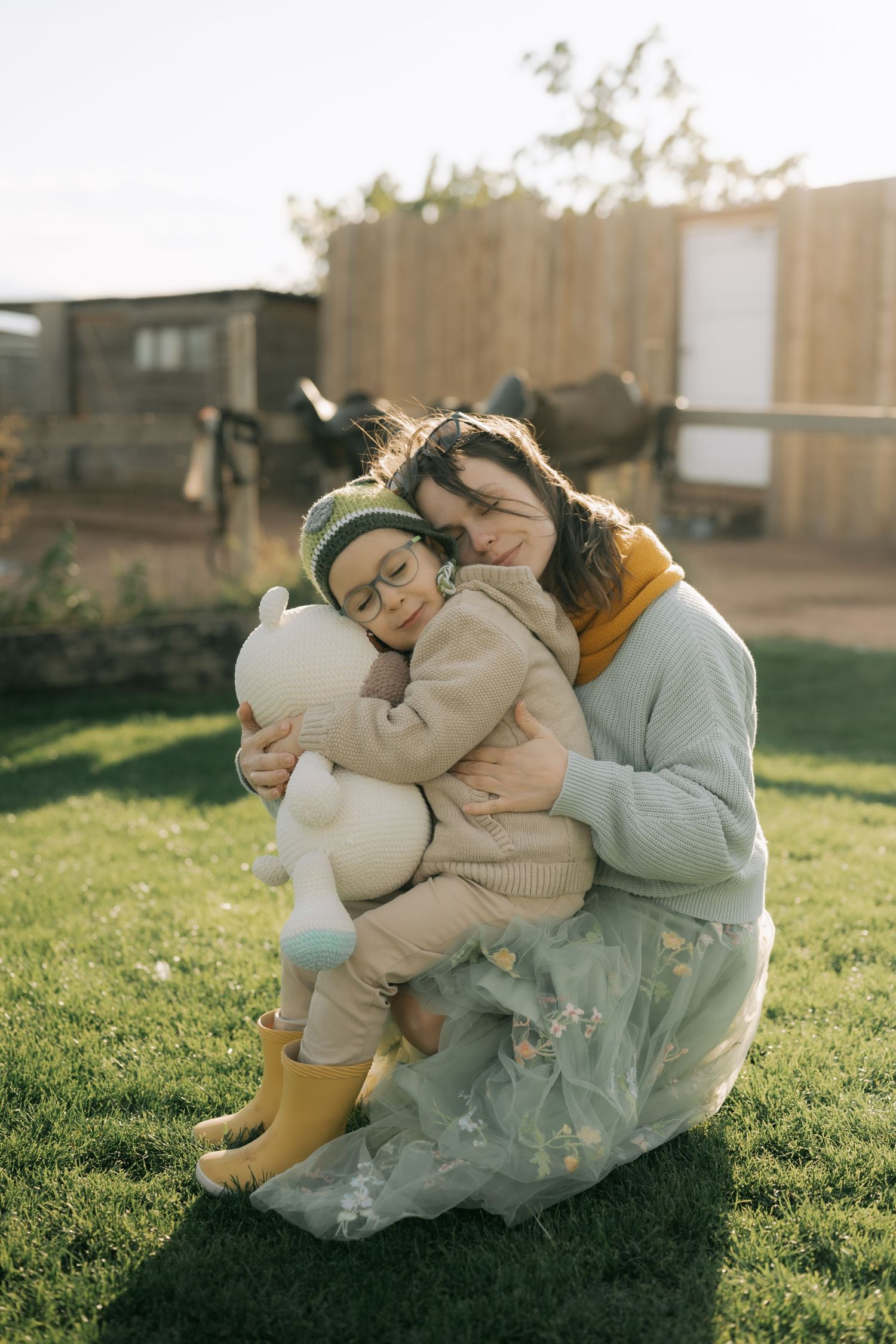 Two people share a tender embrace while sitting on grass in warm sunlight, with one holding a plush toy.