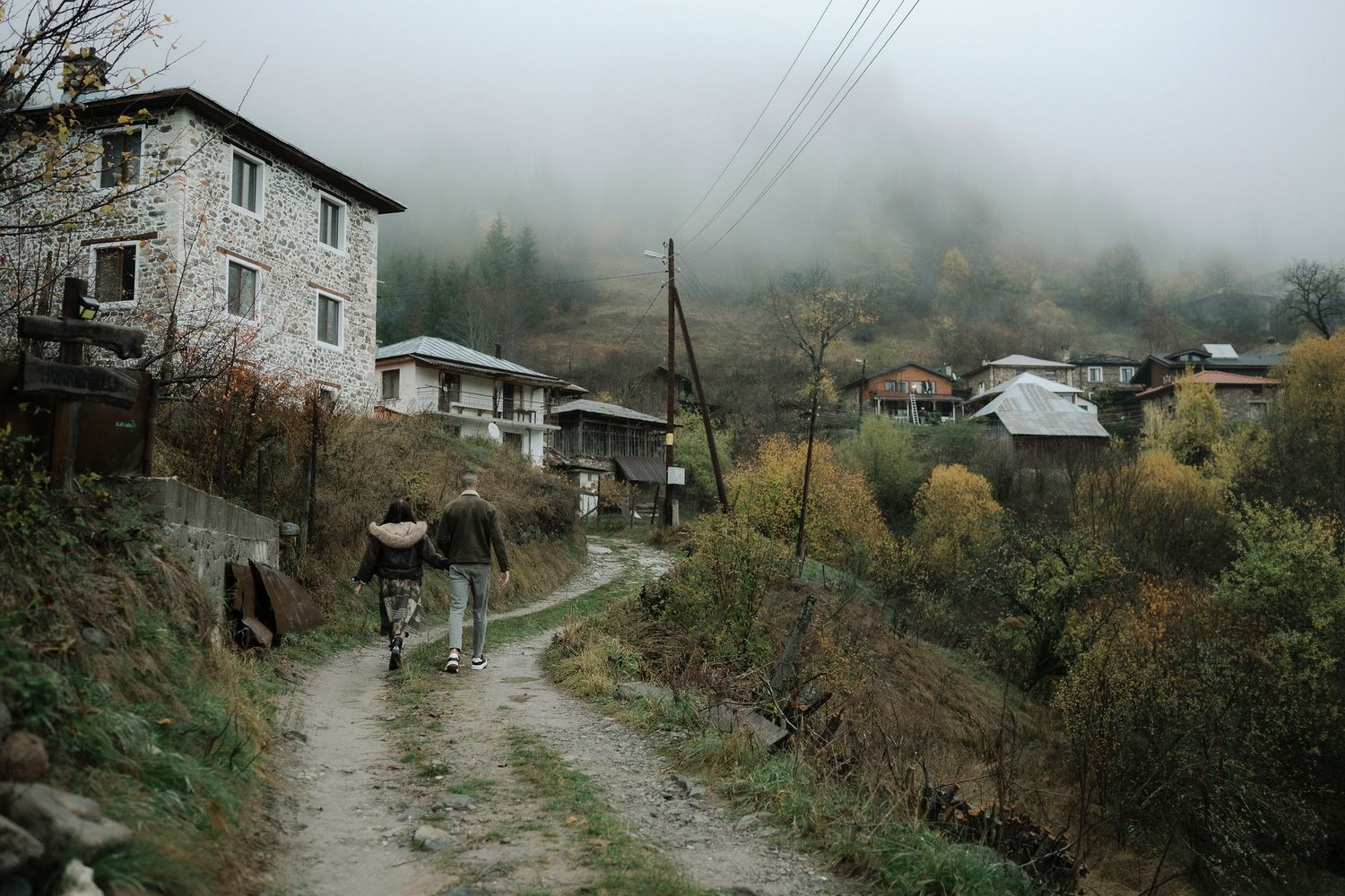 Misty mountain village with stone houses and power lines along a winding dirt path in autumn.